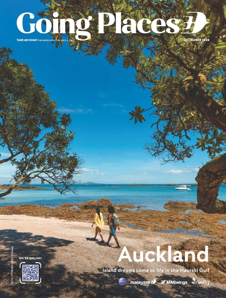 A magazine cover titled 'Going Places' for December 2025, featuring a scenic coastal view of Auckland, New Zealand. The image shows two women walking along a rocky beach, with clear blue water, boats, and lush tree branches framing the scene.