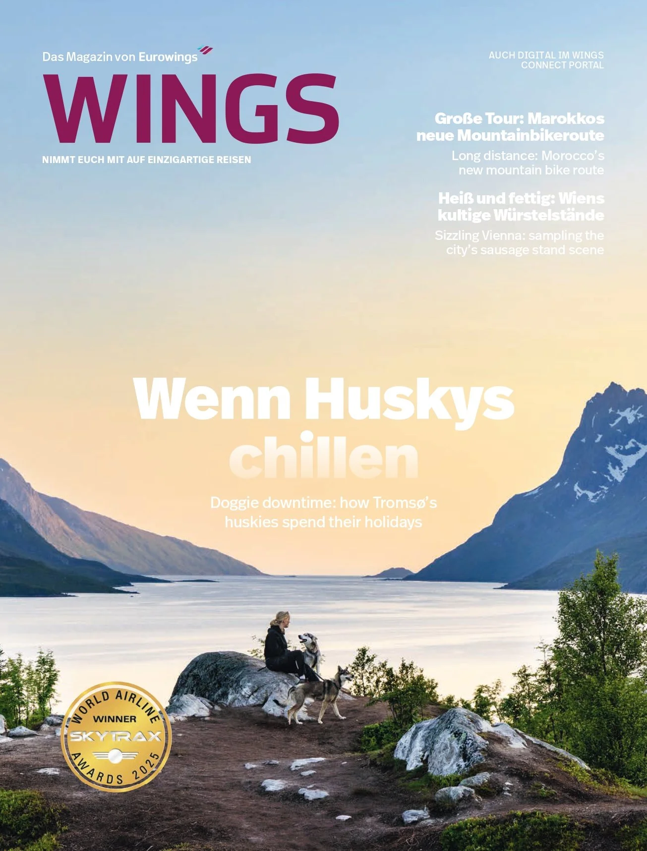 Magazine cover featuring a person sitting with two huskies on a rocky elevation overlooking a fjord with mountains and a sunset sky, promoting travel and dog-friendly destinations.