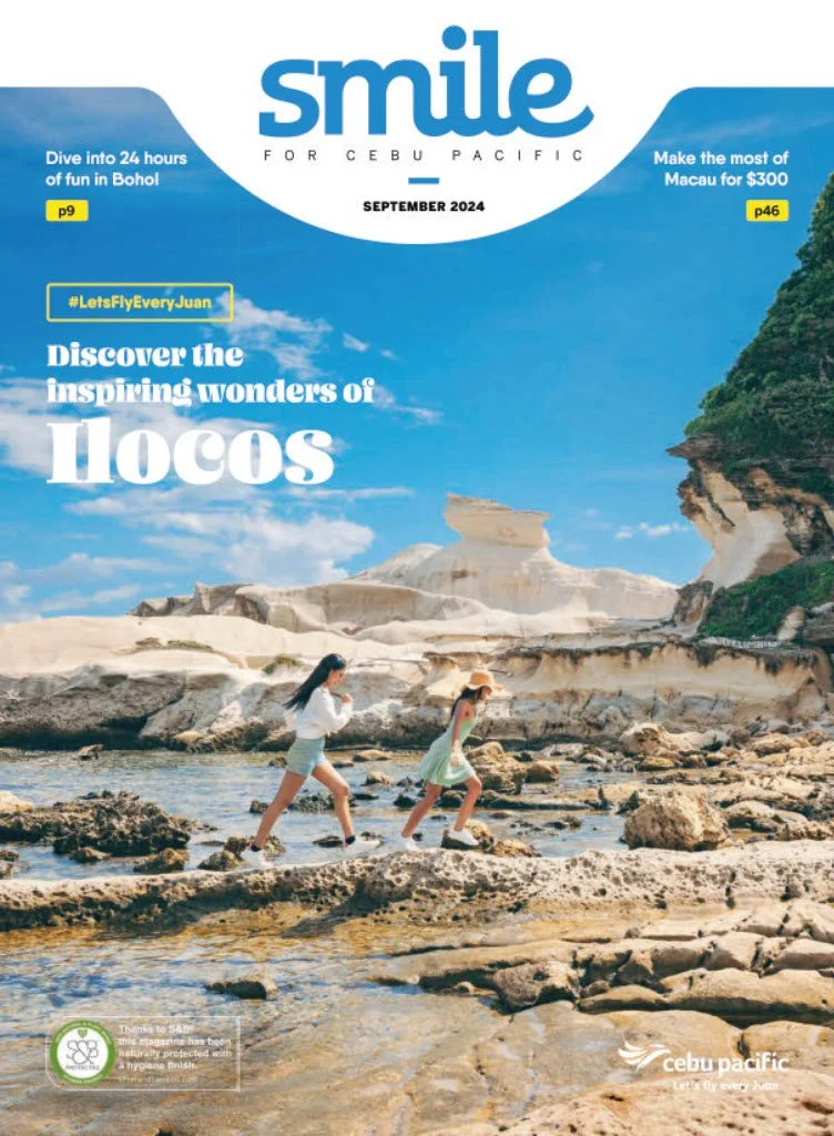Two young women crossing a rocky stream in a scenic coastal landscape with white rock formations, blue sky, and greenery on cliffs in the background.