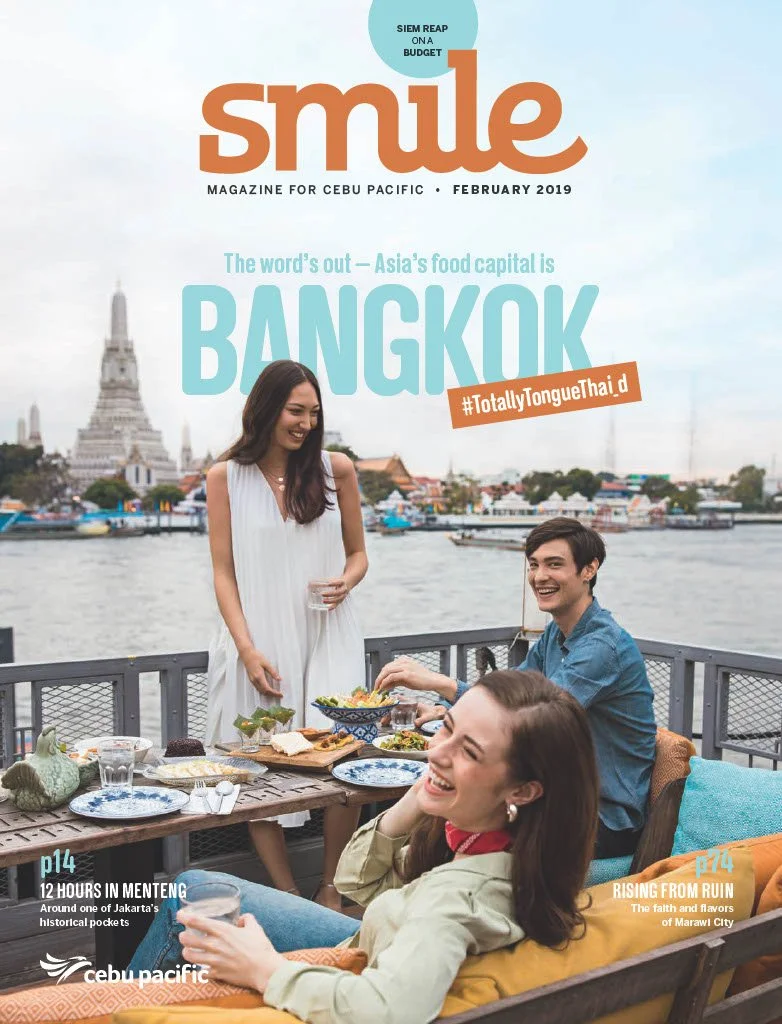 Three friends enjoying a meal outdoors with a river and historic temple in Bangkok in the background on the cover of Smile magazine, February 2019.