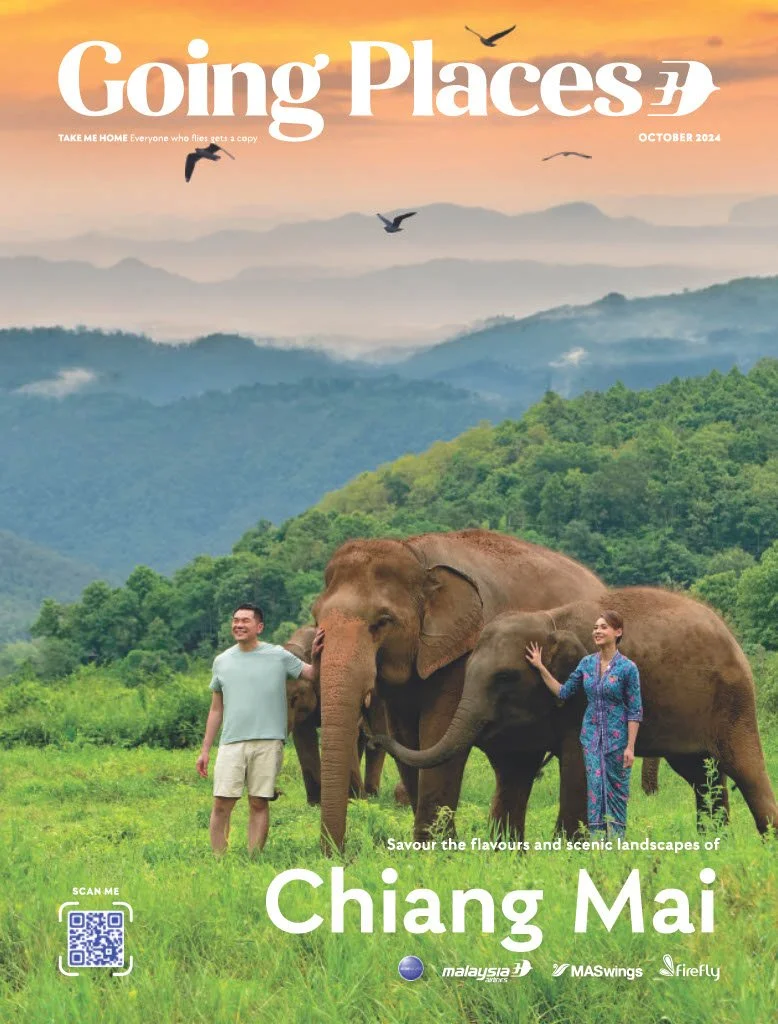A magazine cover featuring a scenic landscape of mountains and green forests in Chiang Mai, Thailand, with three elephants and two tourists, one man and one woman, standing among them. The sky is orange with birds flying overhead, and the magazine is titled "Going Places" October 2024.