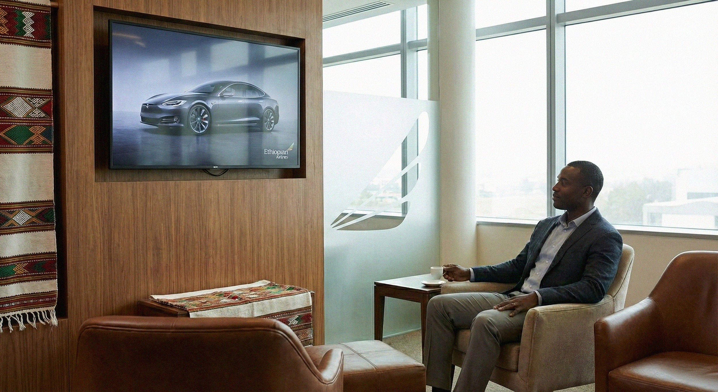 A man in a suit sitting in an office lobby, holding a cup, watching a TV screen showing a silver car with Ethiopian Airlines logo.