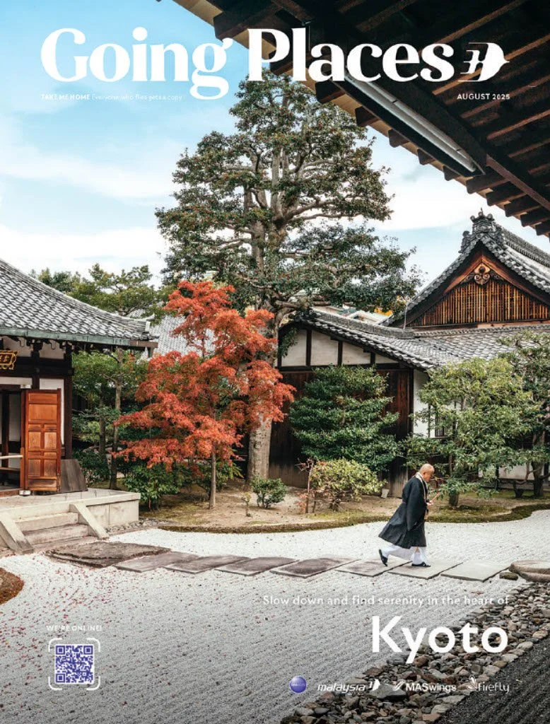 A person dressed in traditional Japanese attire walking on a stone path in a serene Japanese garden with traditional buildings, trees, and autumn foliage, in Kyoto, Japan.