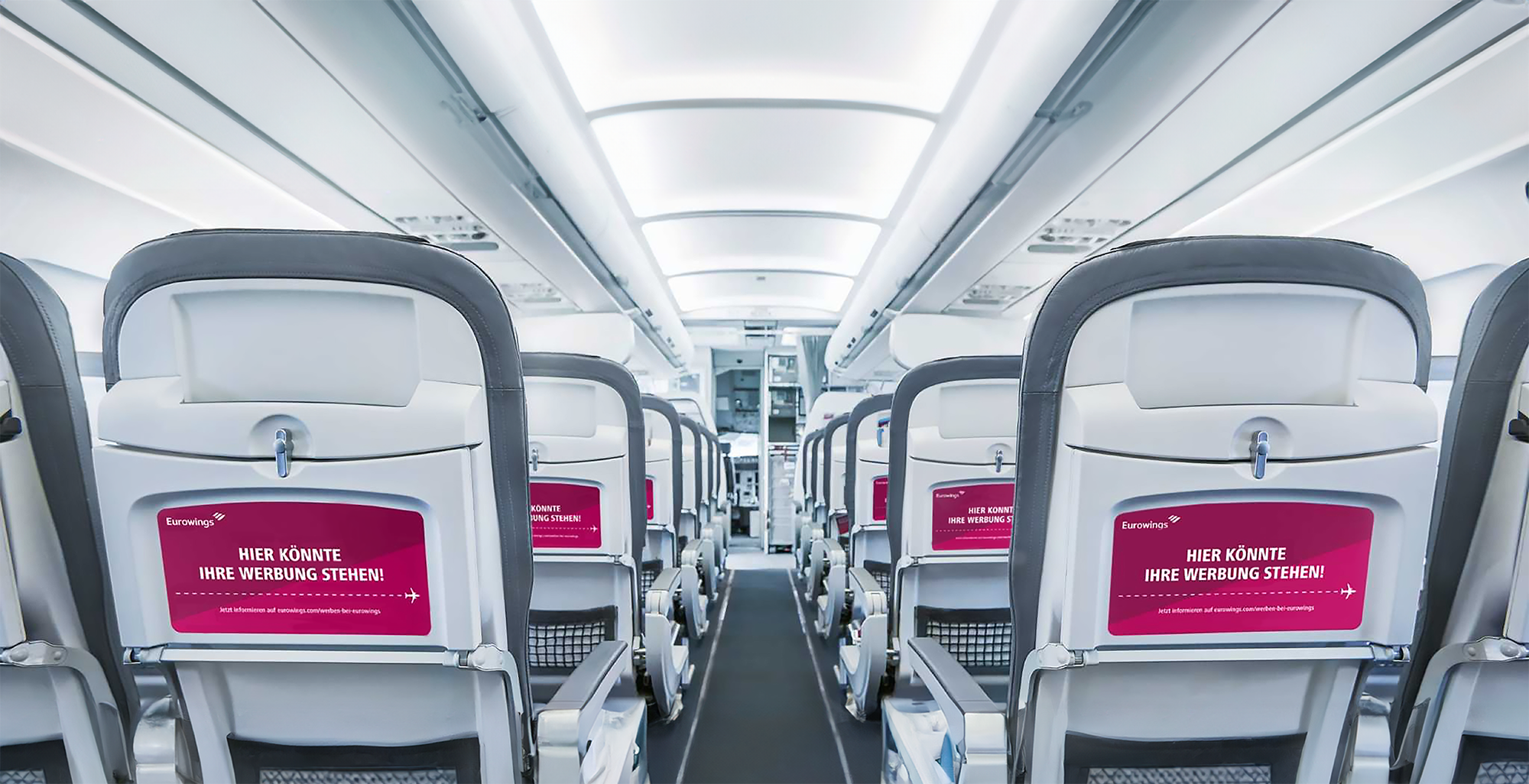 Inside view of an airplane cabin showing rows of white airplane seats with gray headrests. The seatbacks have pink advertisements with German text. The cabin is brightly lit with overhead lighting, and the aisle runs down the middle, leading to the cockpit.