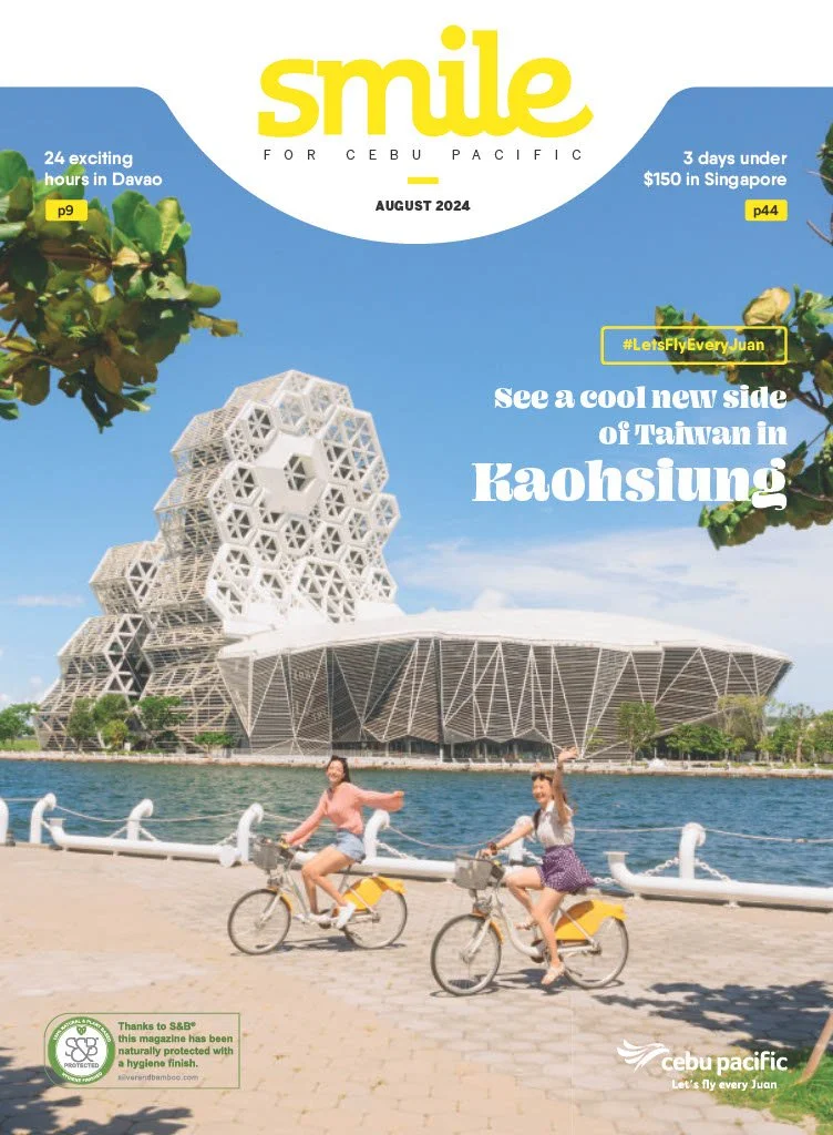 Two women cycling along a waterfront promenade with a modern architectural building in Kaohsiung, Taiwan, in the background during daylight.