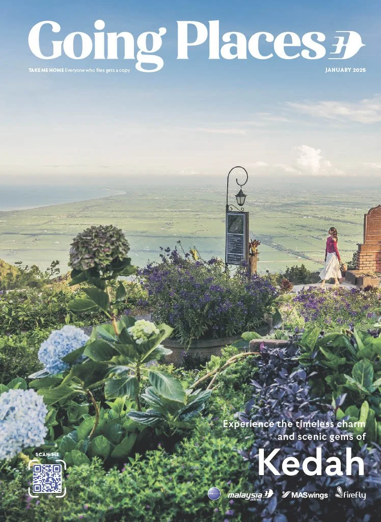 Cover page of Going Places magazine, January 2025 issue, showcasing a scenic landscape from Kedah with lush greenery, flowering plants, a view of the plains and coastline in the background, and a person walking on a path.