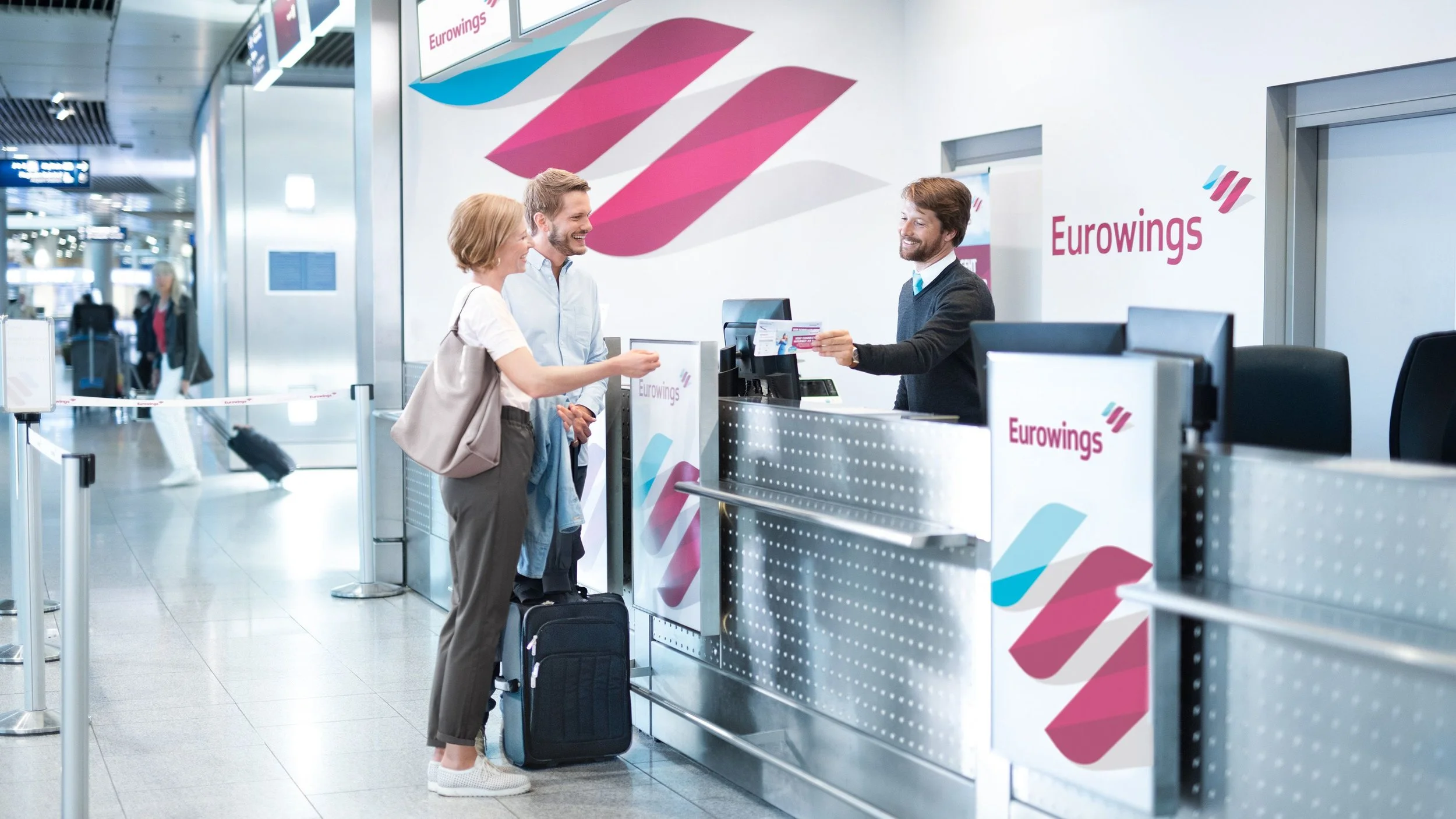 Three travelers at a check-in counter in an airport, exchanging documents with a staff member. The counter displays the Eurowings logo, and other travelers with luggage are visible in the background.