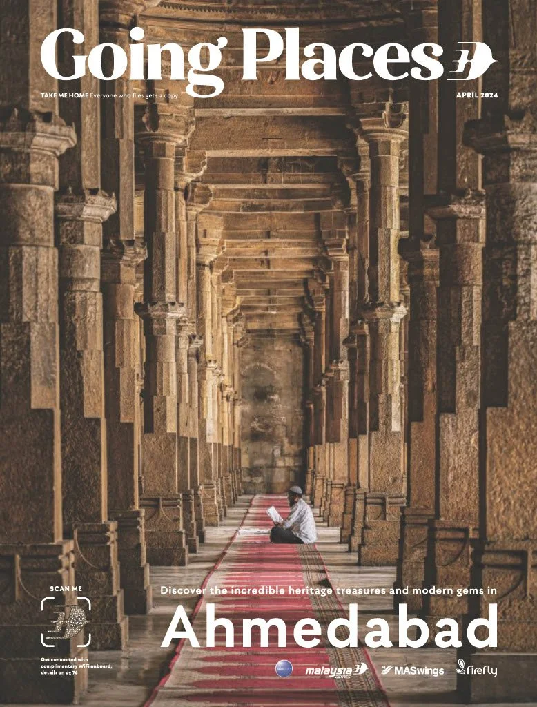 A person sitting on the floor inside an ancient stone corridor, reading a book. The corridor has tall stone columns and a red patterned carpet. The magazine cover mentions Ahmedabad, highlighting heritage treasures and modern gems.