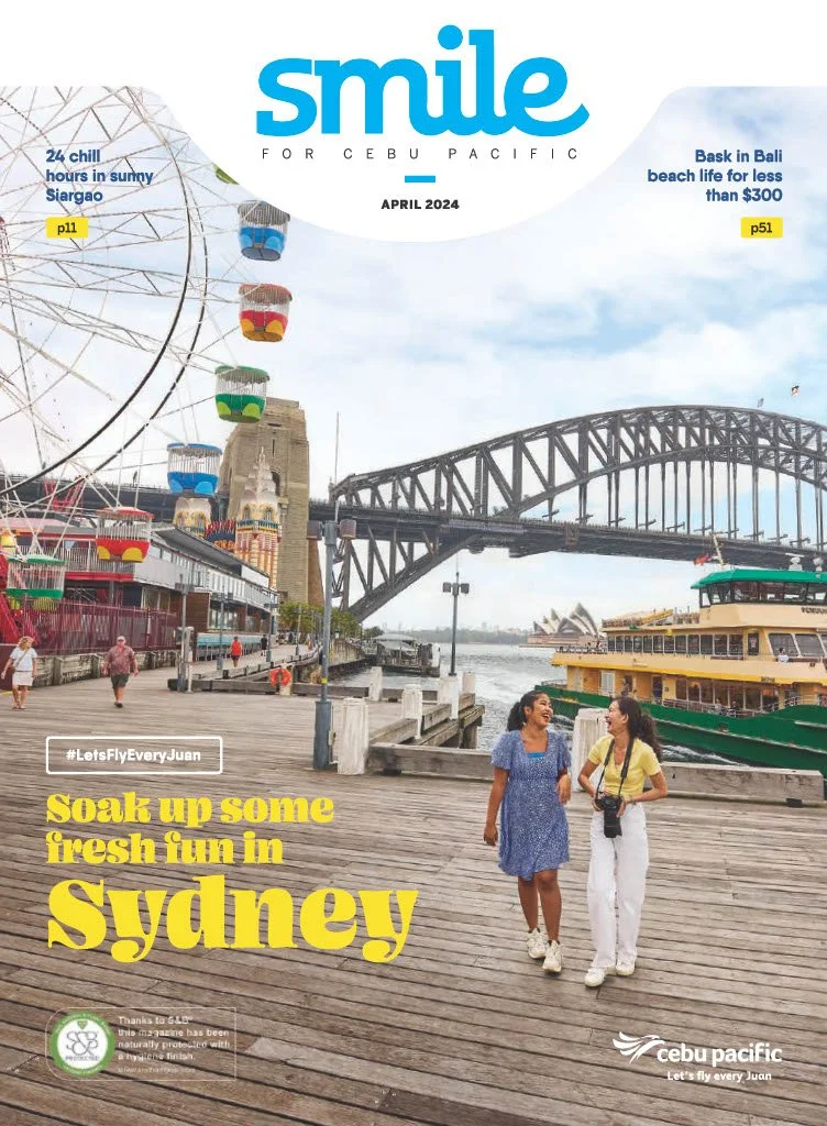 Two women walking and chatting on a wooden pier near the Sydney Harbour with the Harbour Bridge and a ferry in the background, a Ferris wheel, and the Sydney Opera House, during daytime.