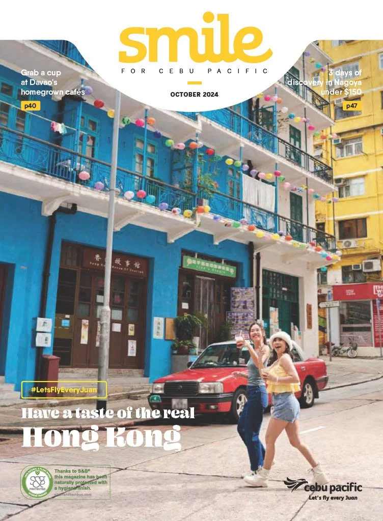 Two women walking on a street in Hong Kong, smiling, with a red car parked in front of a colorful building decorated with balloons, promotional text about travel experiences, and magazine branding.