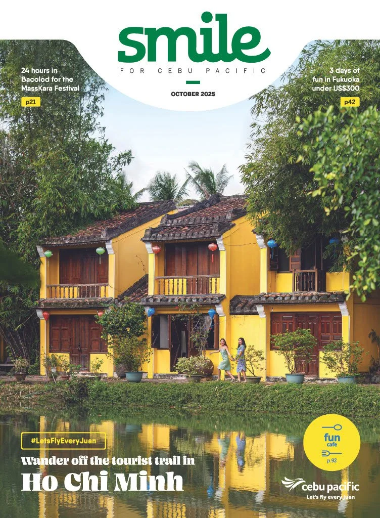 A vibrant yellow two-story house with wooden balcony doors, set beside a calm river with reflections of the house and trees. Two women walk along a path in front of the house, surrounded by lush greenery and potted plants. The sky is partially cloudy.