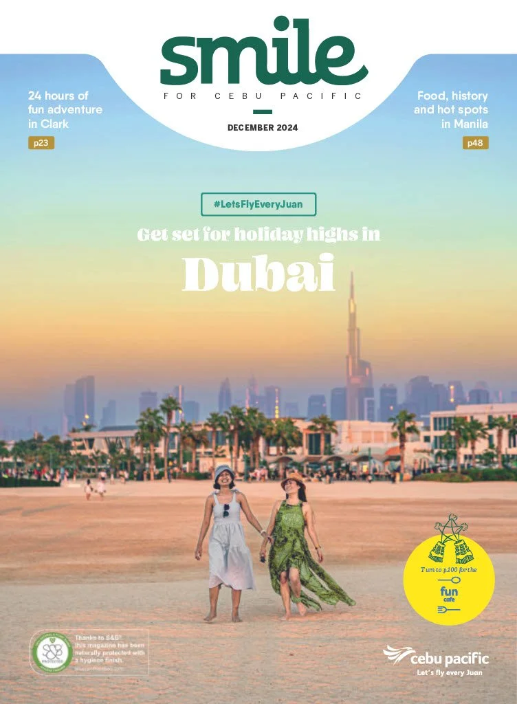 Two women walking on a beach in Dubai with the city skyline, including the Burj Khalifa, in the background, during sunset.