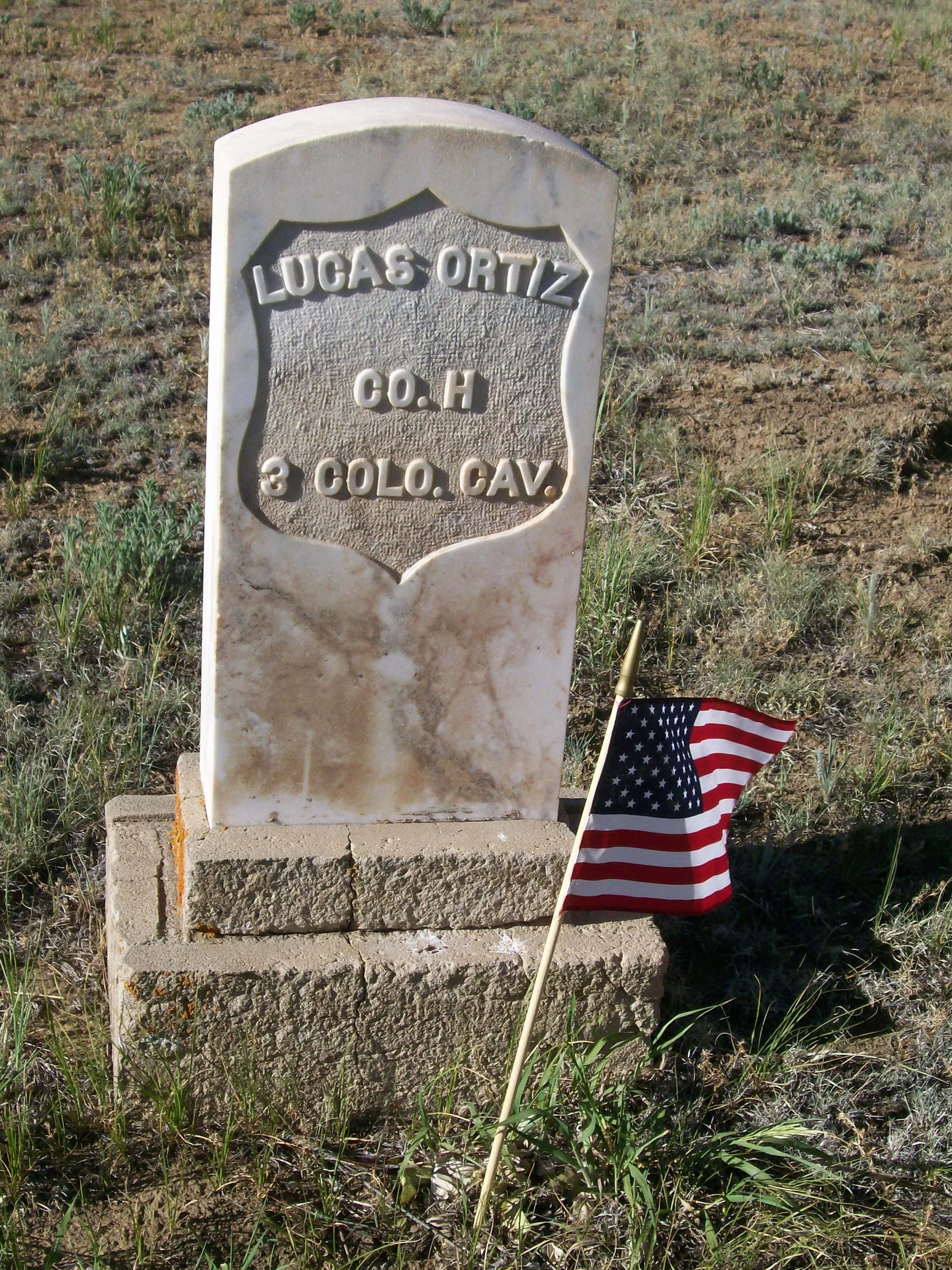 White marble gravestone next to an American Flag. Gravestone reads "Lucas Ortiz, CO.H, 3 COLO. CAV.