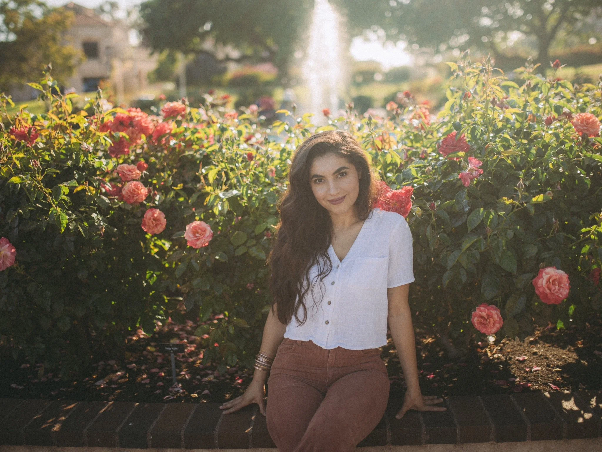 Young woman with long dark hair sitting on a brick ledge in front of a bush with pink roses, in a park or garden during daytime.