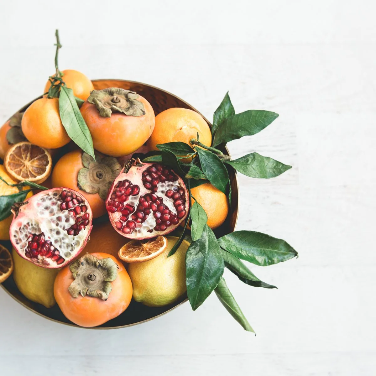 A bowl of persimmons, pomegranates, and dried citrus slices with green leaves on a white surface.