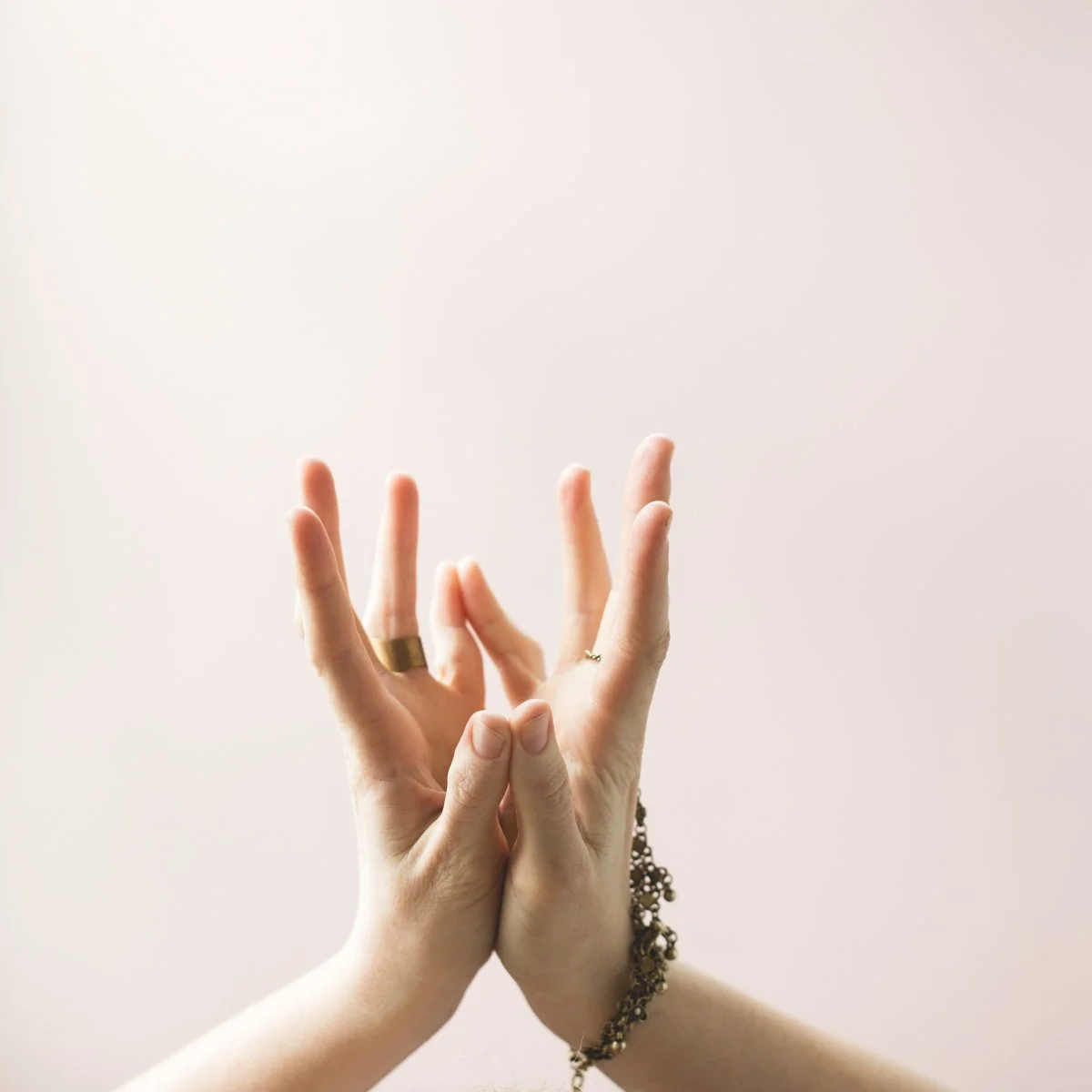 Two hands with rings and a bracelet are making a mudra gesture against a plain, light-colored background.