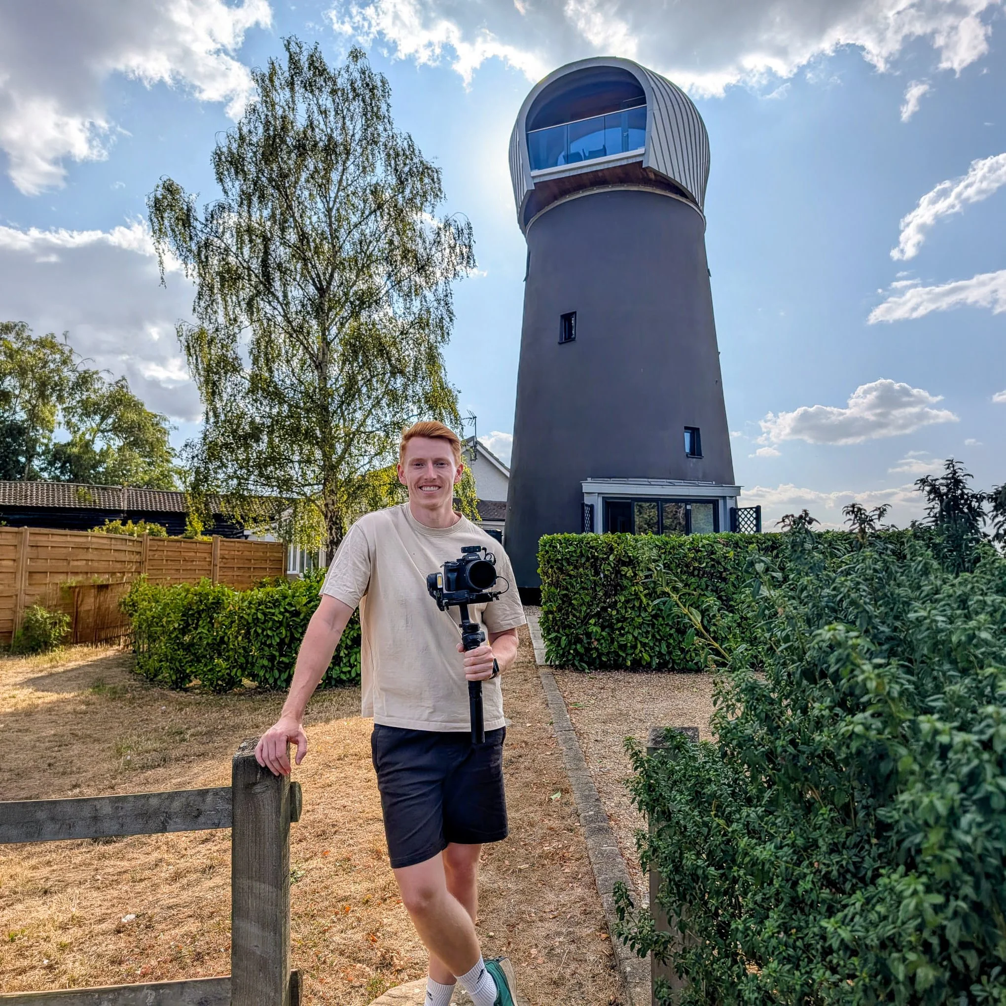 Man holding a camera stabilizer beside a modern converted windmill tower in a sunny garden