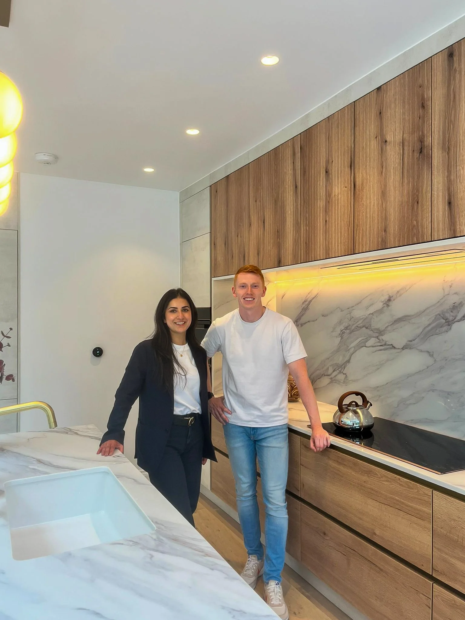 Two people, a woman and a man, standing in a modern kitchen with wooden cabinets, marble backsplash, and a white marble countertop. The woman has long dark hair and is wearing a dark blazer over a white shirt, while the man has short red hair and is wearing a white t-shirt and jeans. They are smiling and posing for the photo.