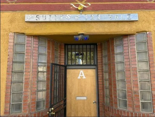 Entrance to a building with brick walls, a wooden door with the letter 'A' on it, and a sign above that reads 'Stuffs at the Vale'.