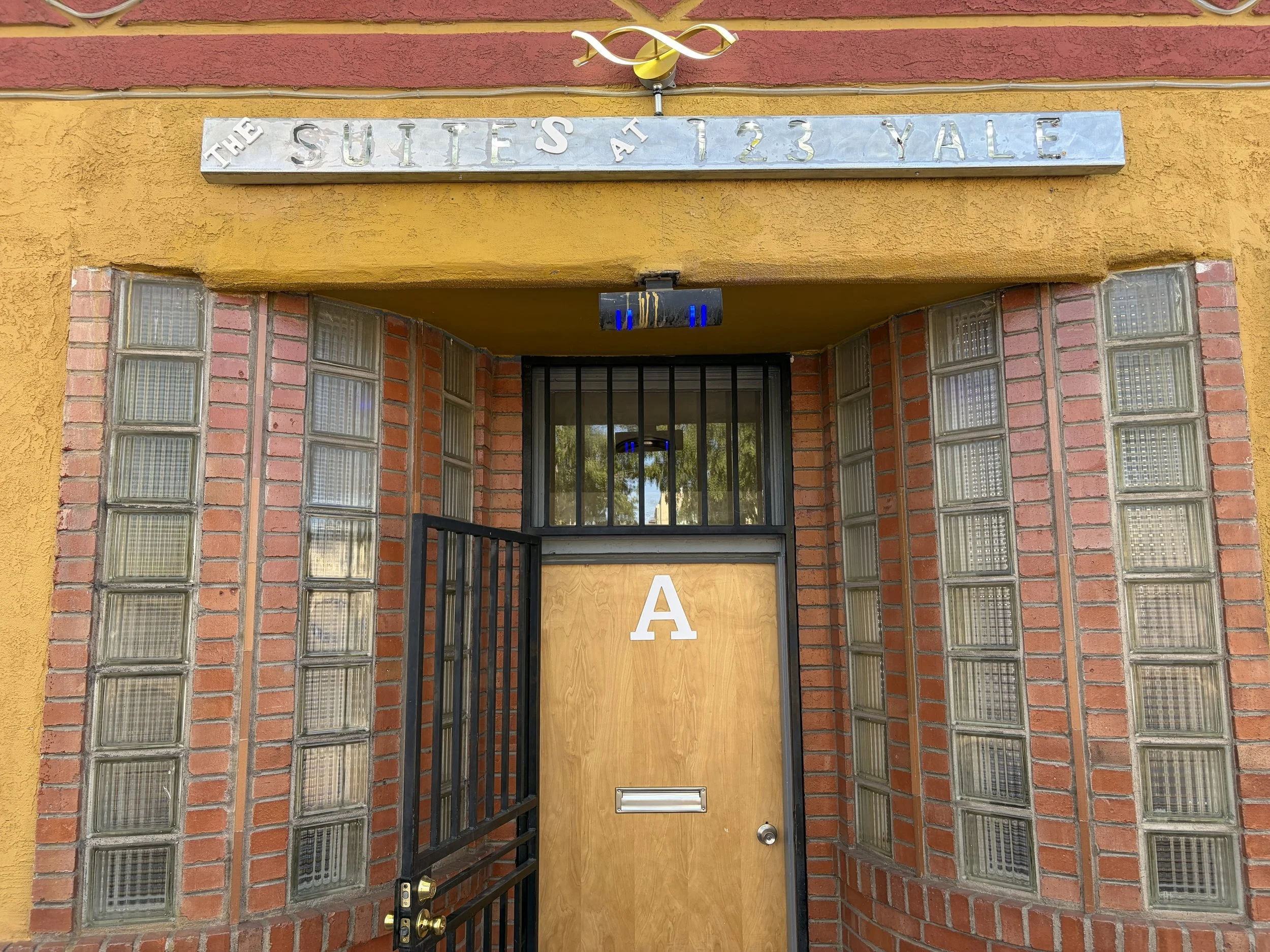 The entrance to a building with a sign that reads 'The Suites at 123 Yale' on a metal plaque above the door. The door has a gate and a mailbox attached to it. The building has a yellow and red exterior with glass block windows on either side of the door.