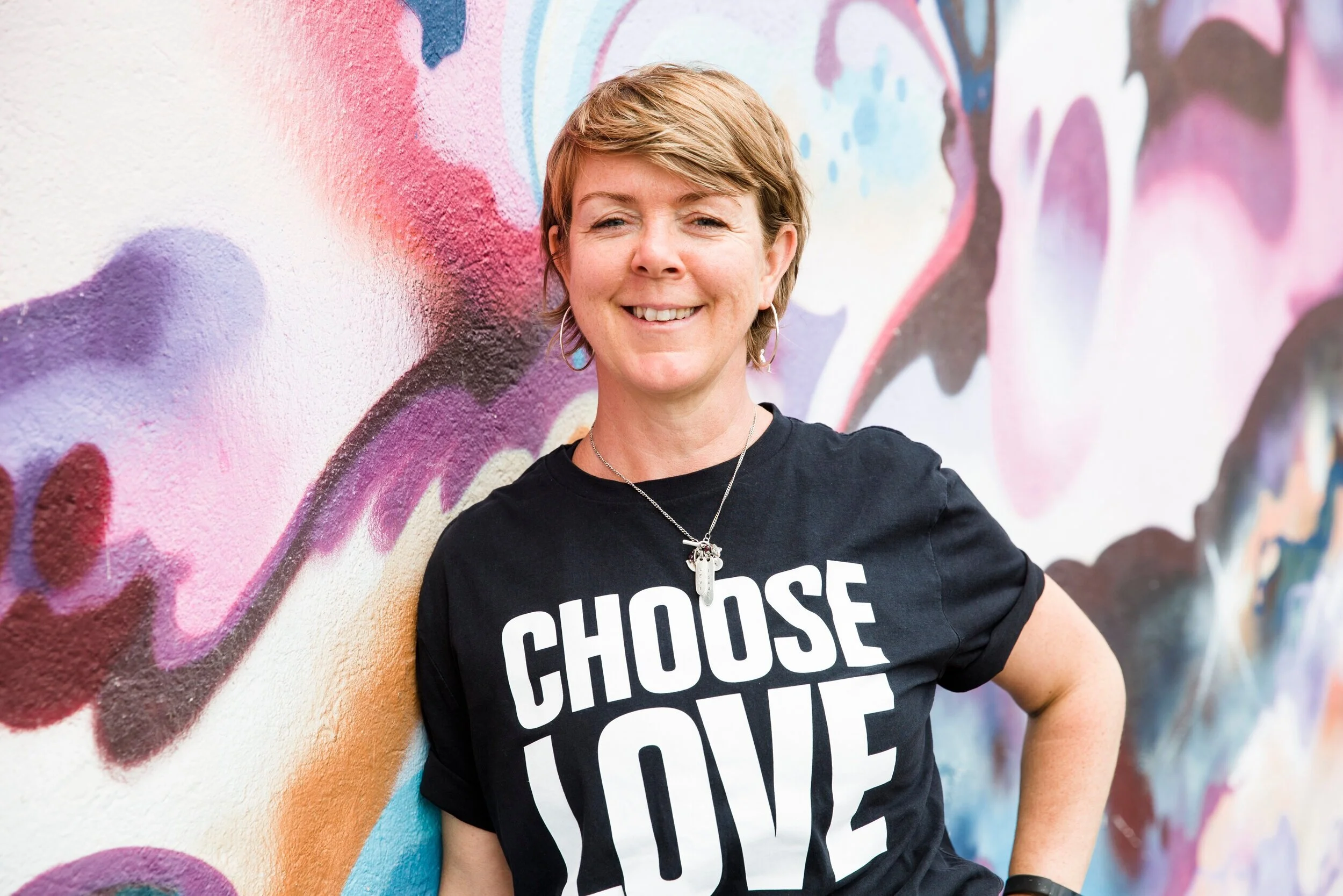 A woman with short blonde hair smiling, wearing a black t-shirt with white text and a necklace with a key pendant, standing against a colorful graffiti wall.