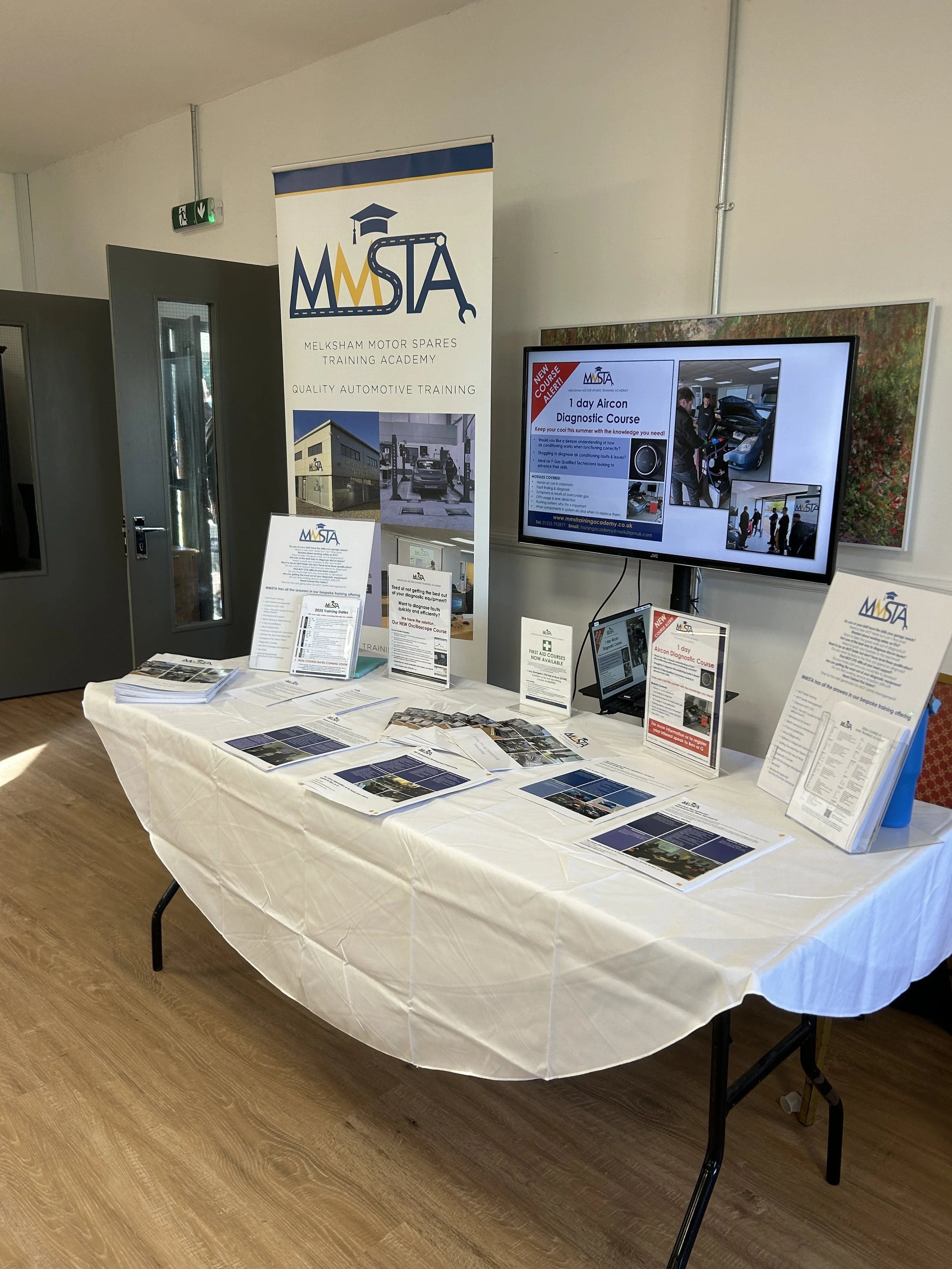 A table with brochures and flyers related to Melksham Motor Spares Training Academy, with a large banner, a monitor displaying course information, and a tablet on display.