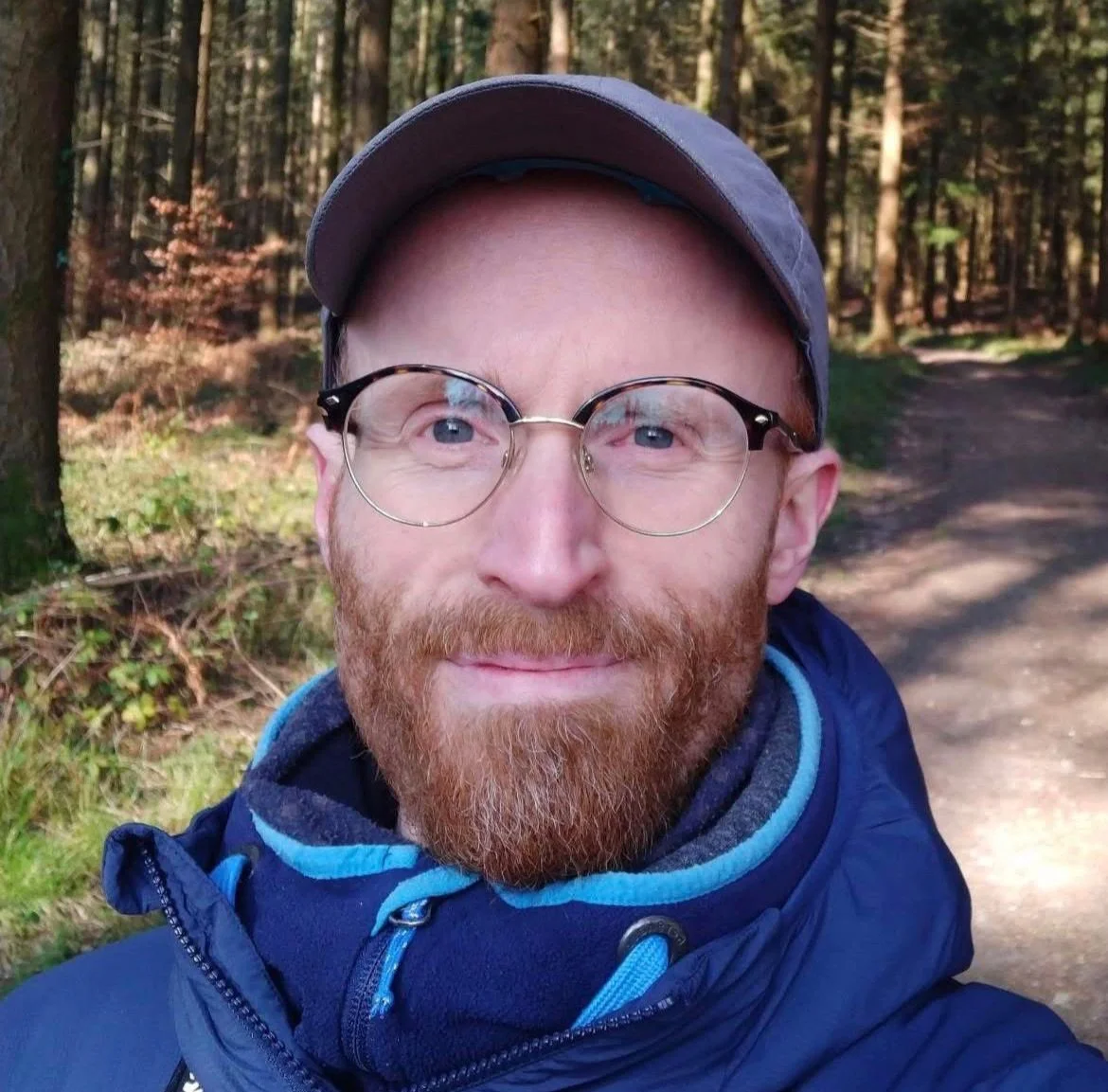 A man with a red beard and glasses taking a selfie outdoors in a wooded area, wearing a baseball cap and a blue jacket.