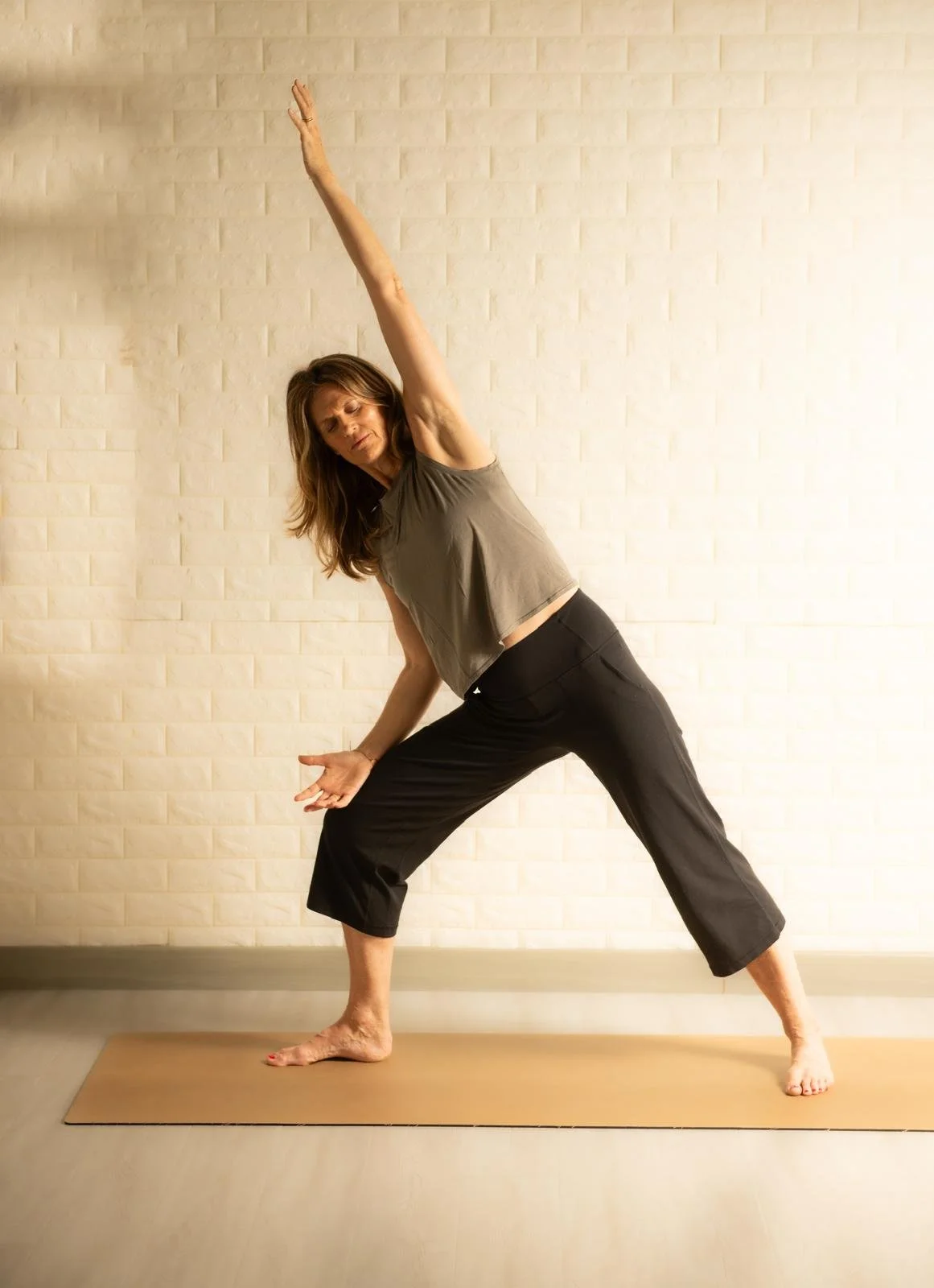 A woman practicing yoga indoors on a beige mat, standing in a wide-legged side bend pose with one arm raised above her head and the other reaching down towards her foot, in front of a light-colored brick wall.