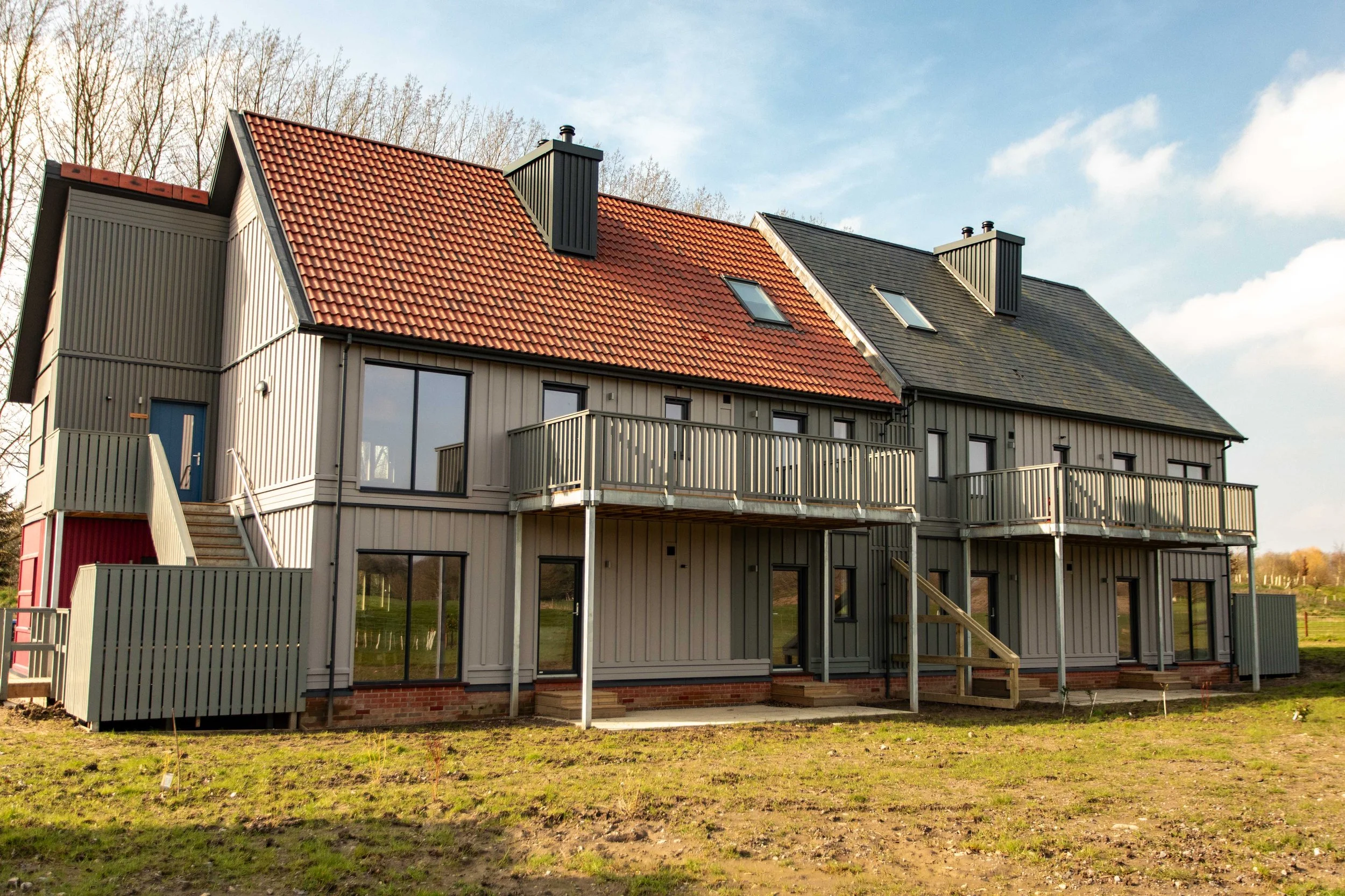 A multi-story residential building with a brick foundation, vertical siding, and a mix of red and dark gray roof shingles. It features multiple balconies, windows, and a staircase leading to an entrance.