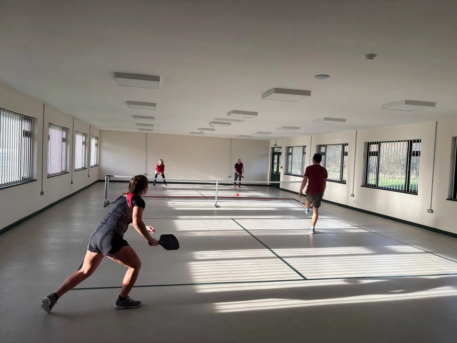 Four people playing pickleball inside an indoor court. The court has a net in the middle and multiple windows along the walls. Two players are on each side of the net, engaging in the game.