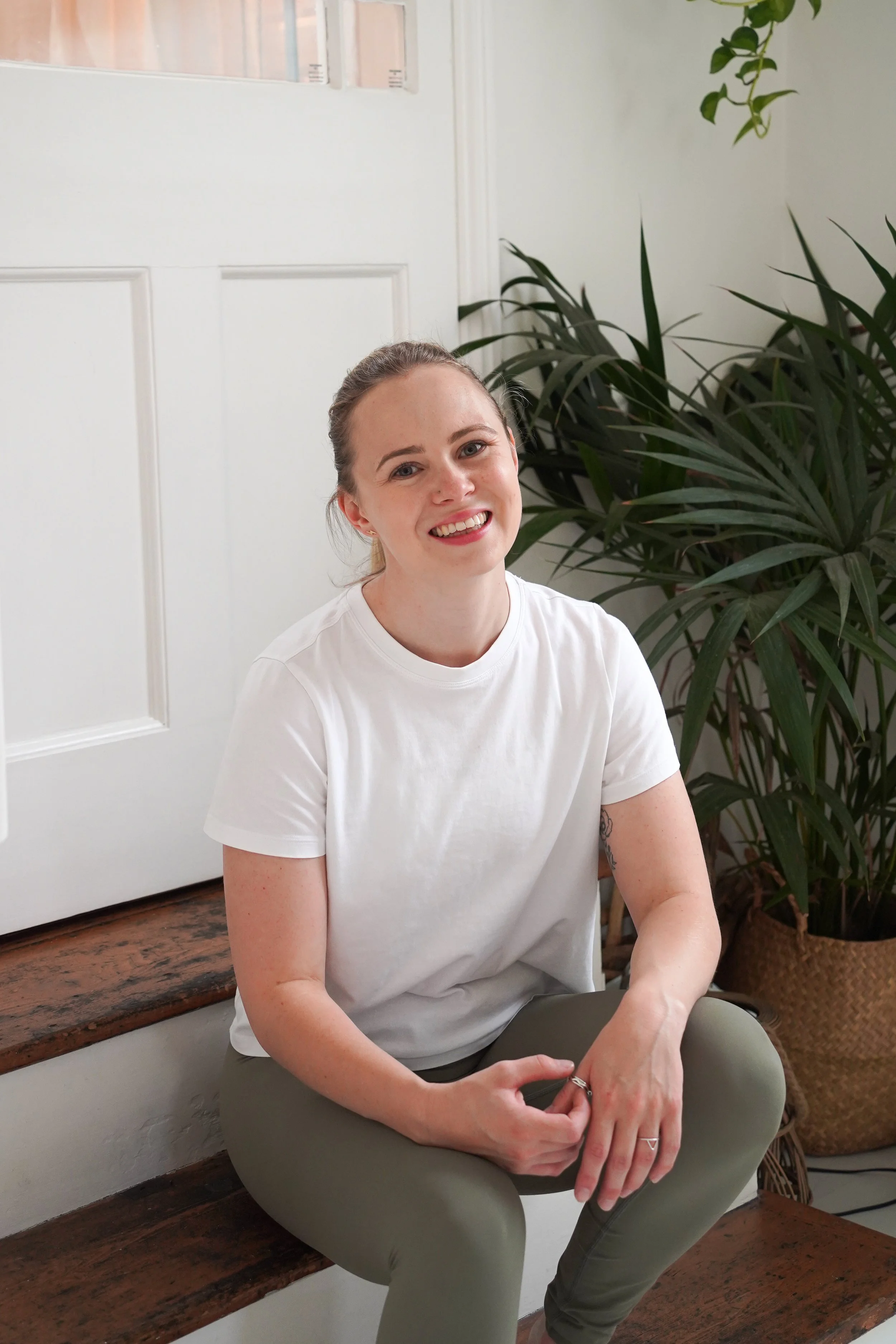 A young woman with light skin, blonde hair tied back, wearing a white t-shirt and gray pants, sitting on a wooden bench indoors. She is smiling and has a tattoo on her left forearm. Behind her, there is a large green leafy plant and a white door with a window.