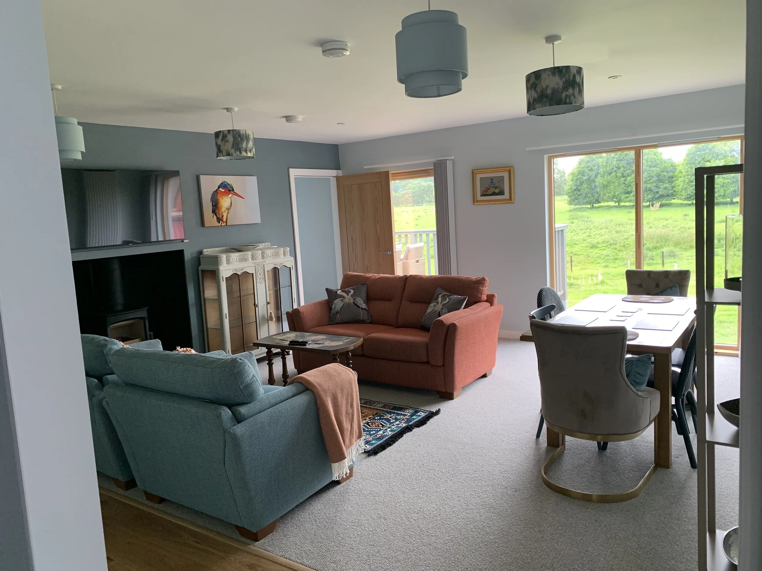 Living room with blue and orange sofas, a white cabinet, and a dining table with six chairs, large windows with a view of a green outdoor landscape.