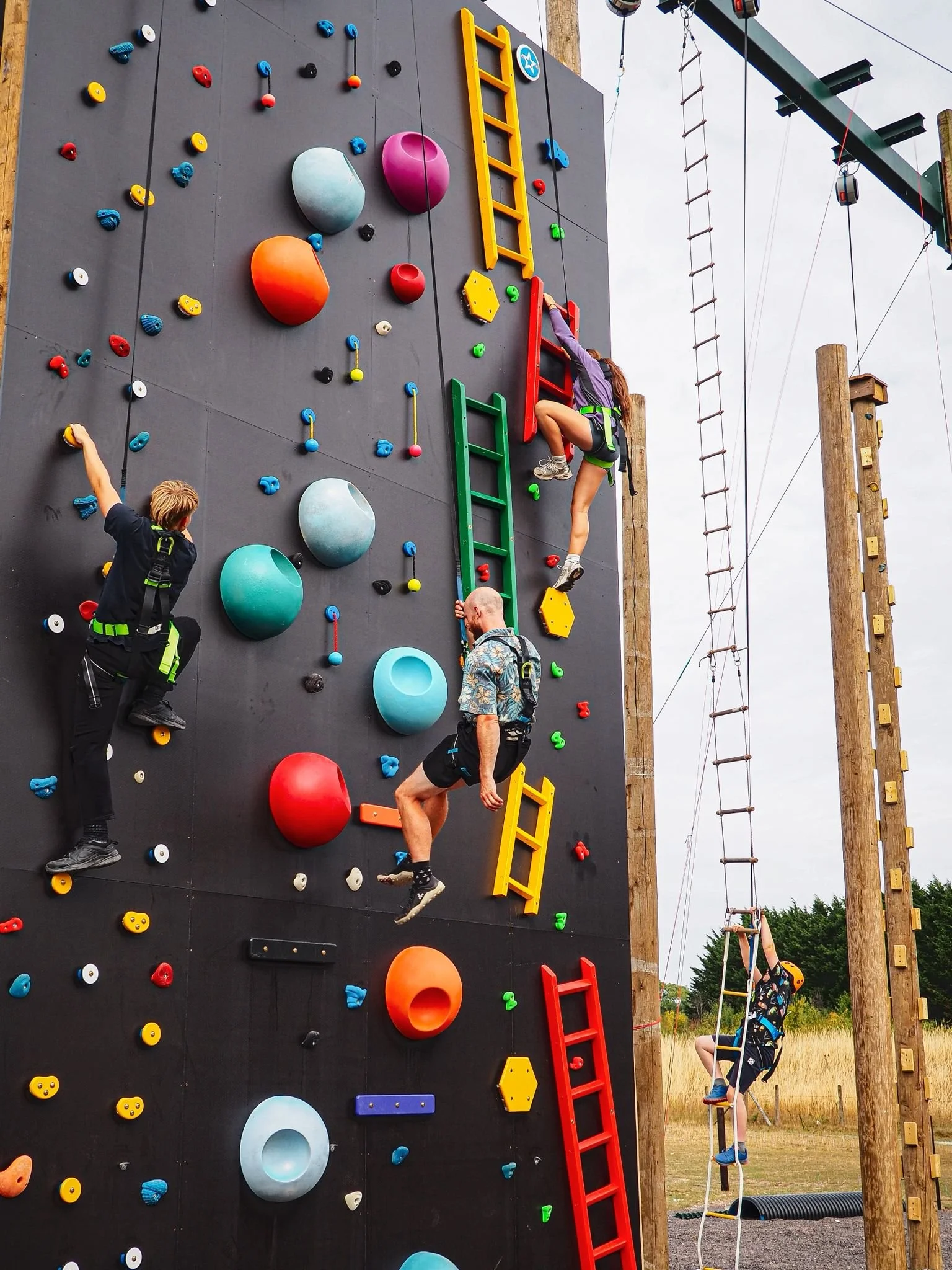 People climbing an outdoor rock climbing wall with colorful holds and ladders, with a safety harness on each person.