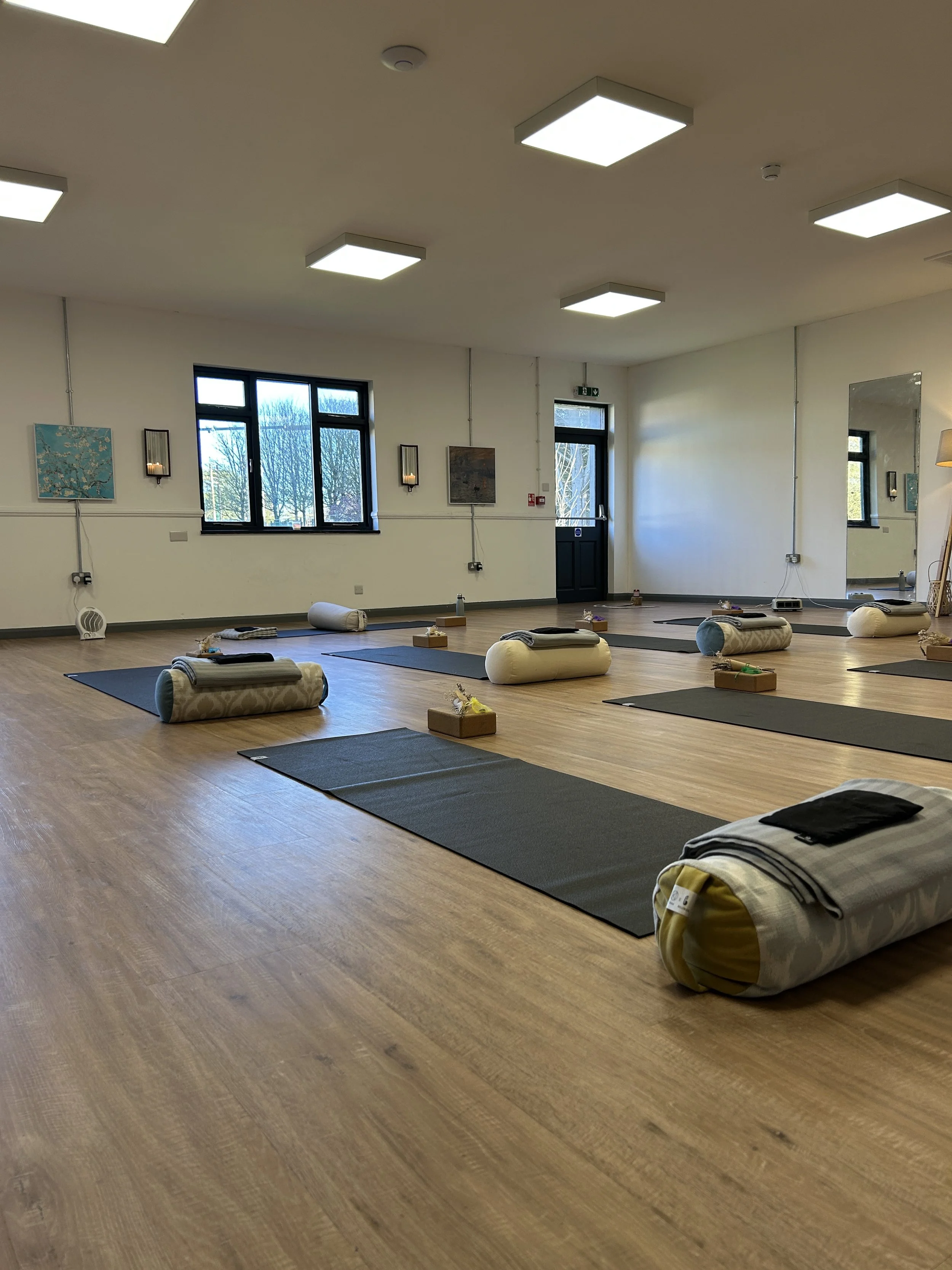 An empty yoga or meditation studio with mats, bolsters, and small wooden blocks arranged on a wooden floor. Large windows let in natural light, and there are paintings on the walls. The room appears to be prepared for a class or session.