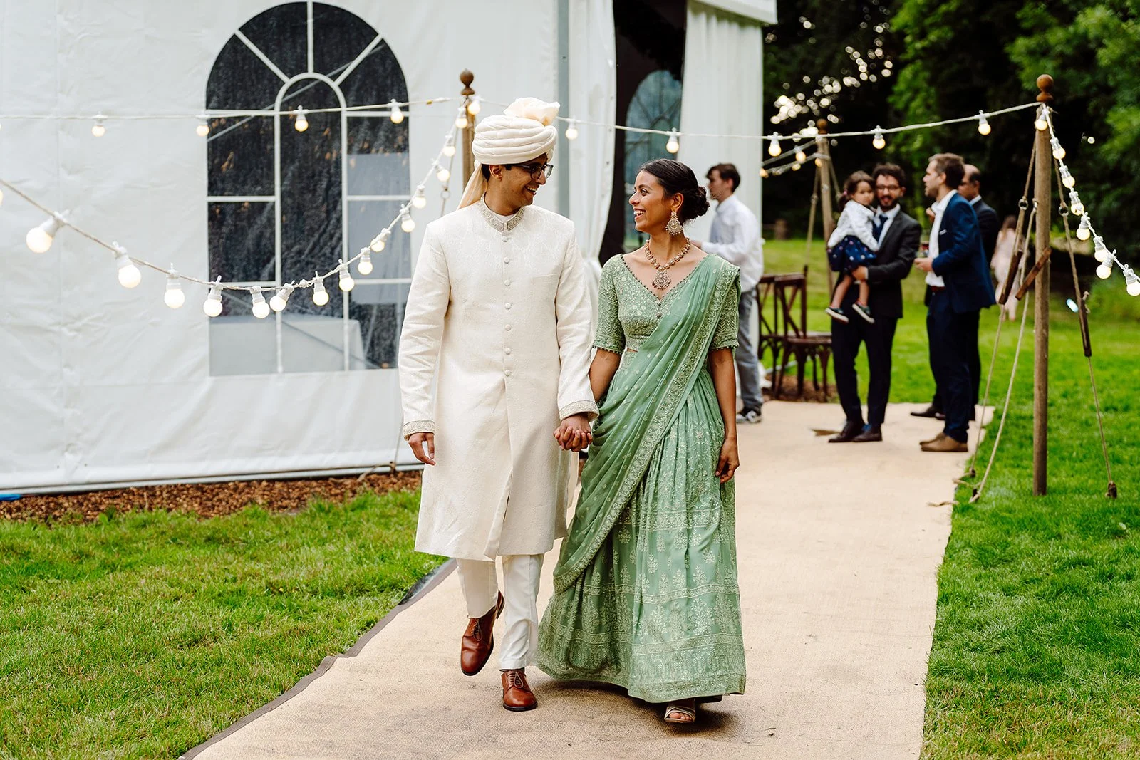 A couple dressed in traditional Indian wedding attire holding hands, walking on a path at an outdoor event with string lights and guests in the background.