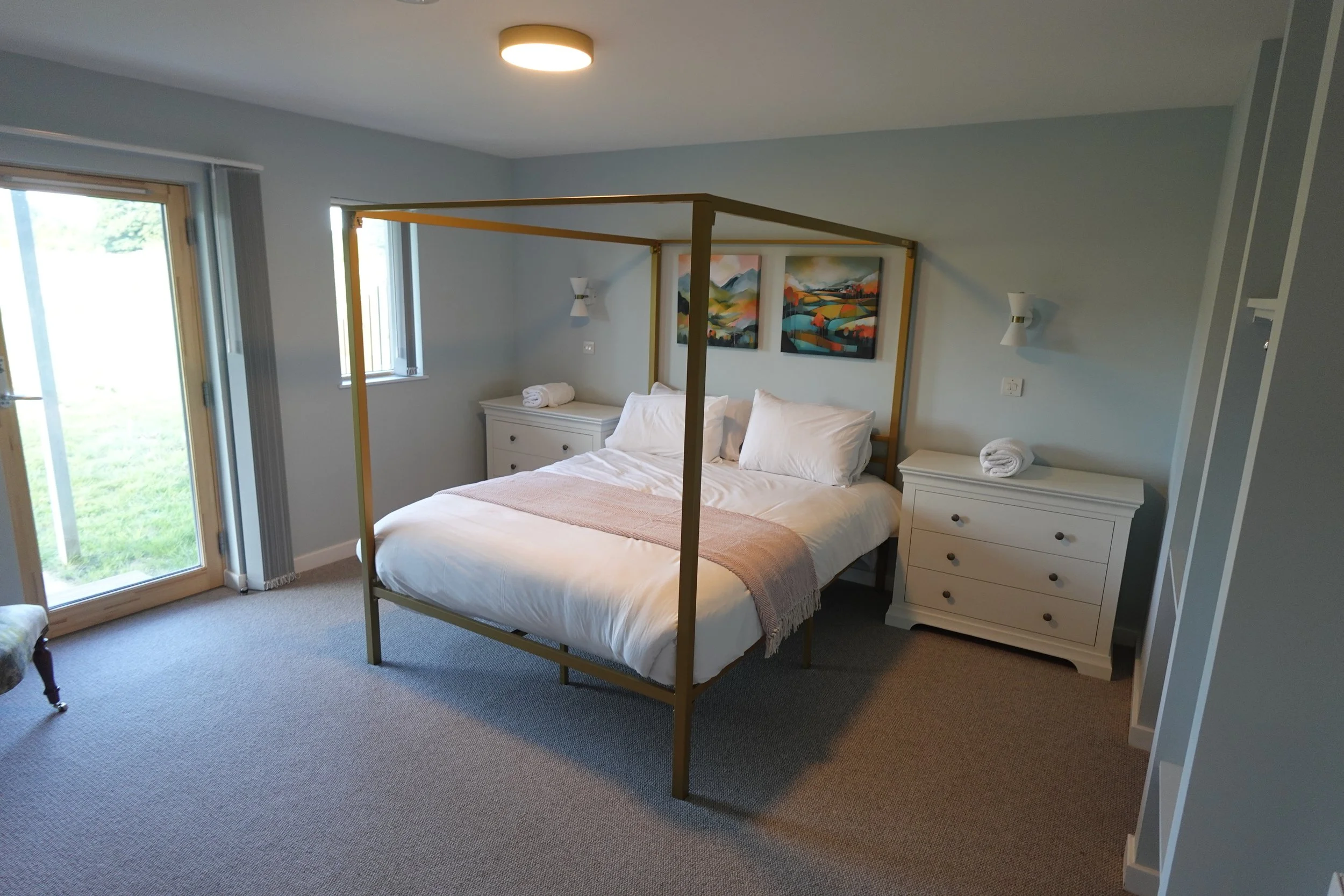 A bedroom with a modern four-poster bed, white bedding, and two white dressers on either side. There are three colorful landscape paintings on the wall above the bed, and a small window and glass door lead to an outdoor area. The room has soft blue walls and neutral carpeting.