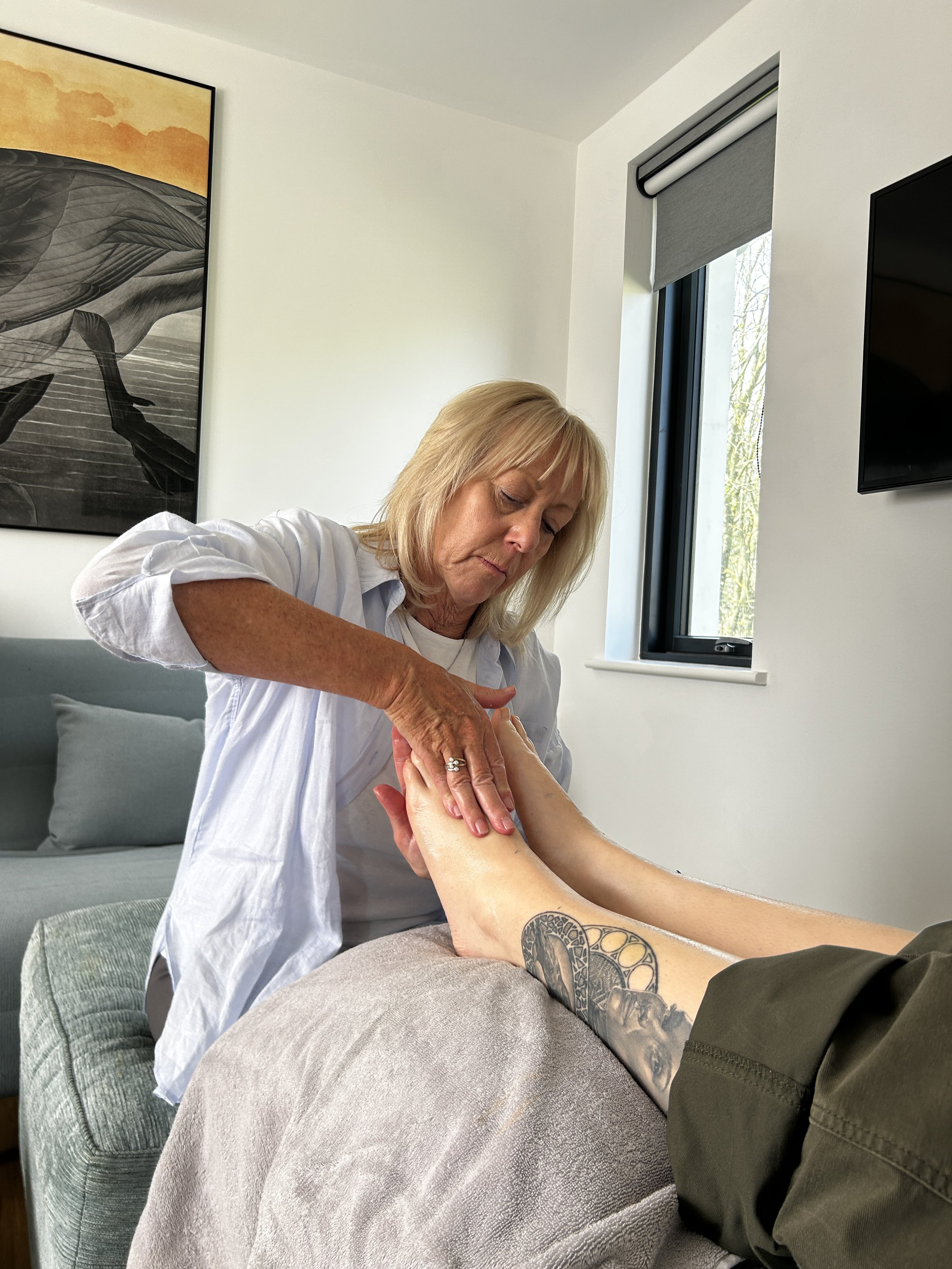 A woman with blonde hair appears to be giving a massage to a person's arm with tattooed forearm while sitting on a bed in a room with white walls, large window, and wall art.