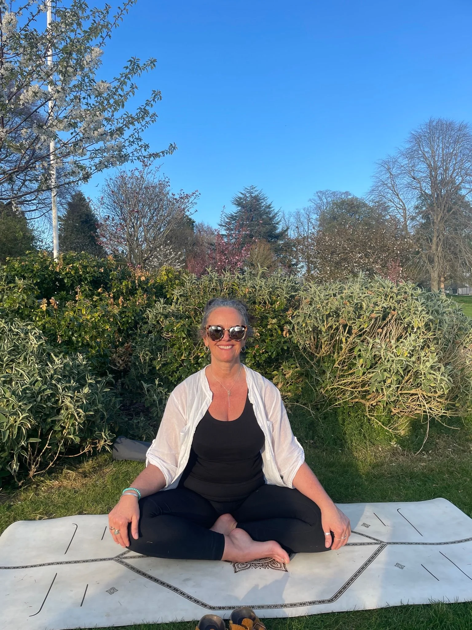 A woman sitting cross-legged on a yoga mat outdoors, smiling, wearing sunglasses, a white jacket over a black top, and black pants. She is in front of lush green bushes with trees and a clear blue sky in the background.