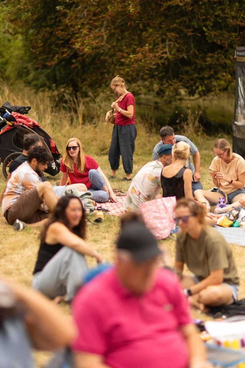 A group of people enjoying a picnic outdoors on a grassy area near a body of water, with some sitting on blankets and others standing.