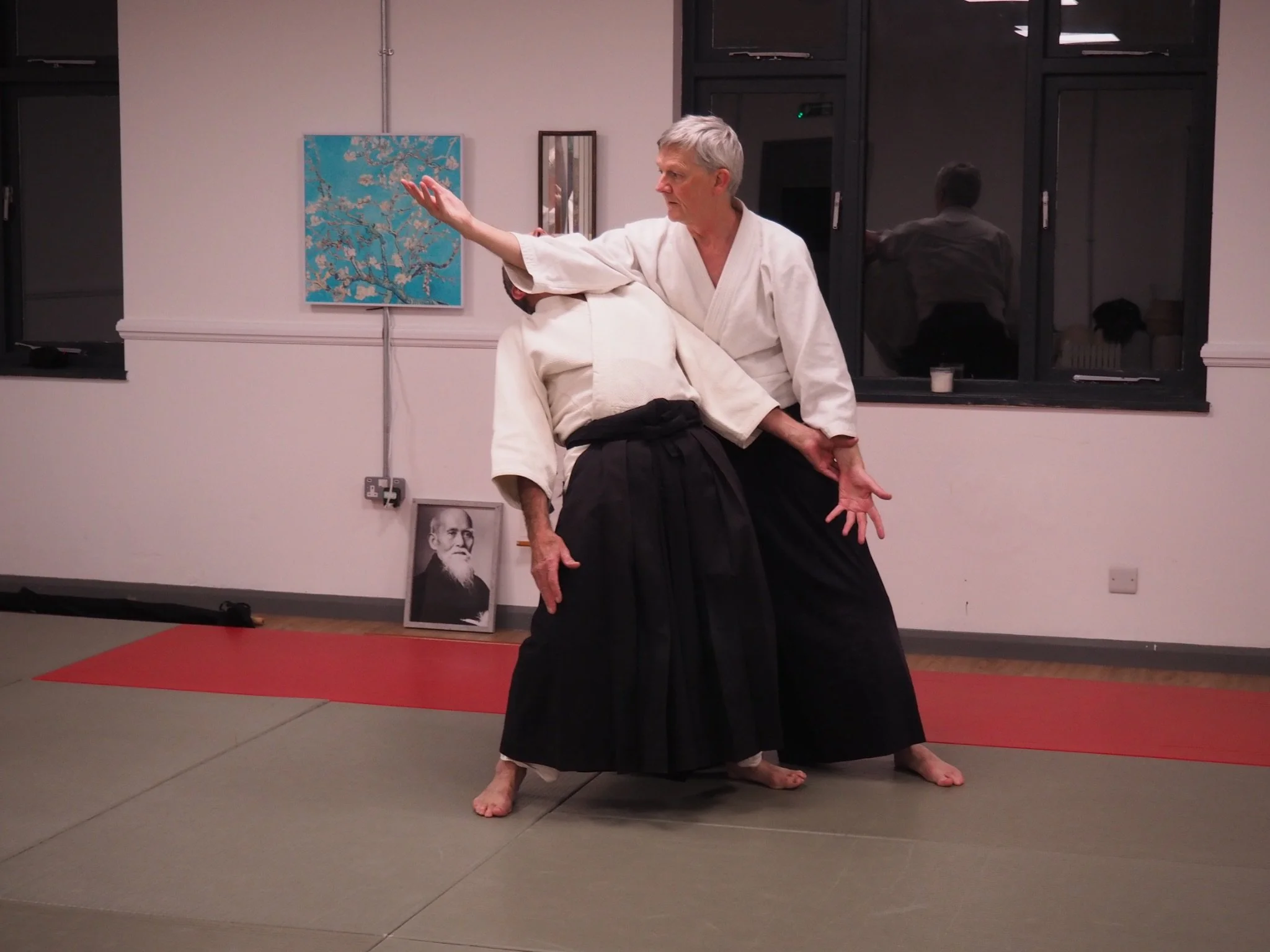 An elderly person practicing martial arts in traditional attire, performing a move with a partner in a room with artwork and photographs on the wall.
