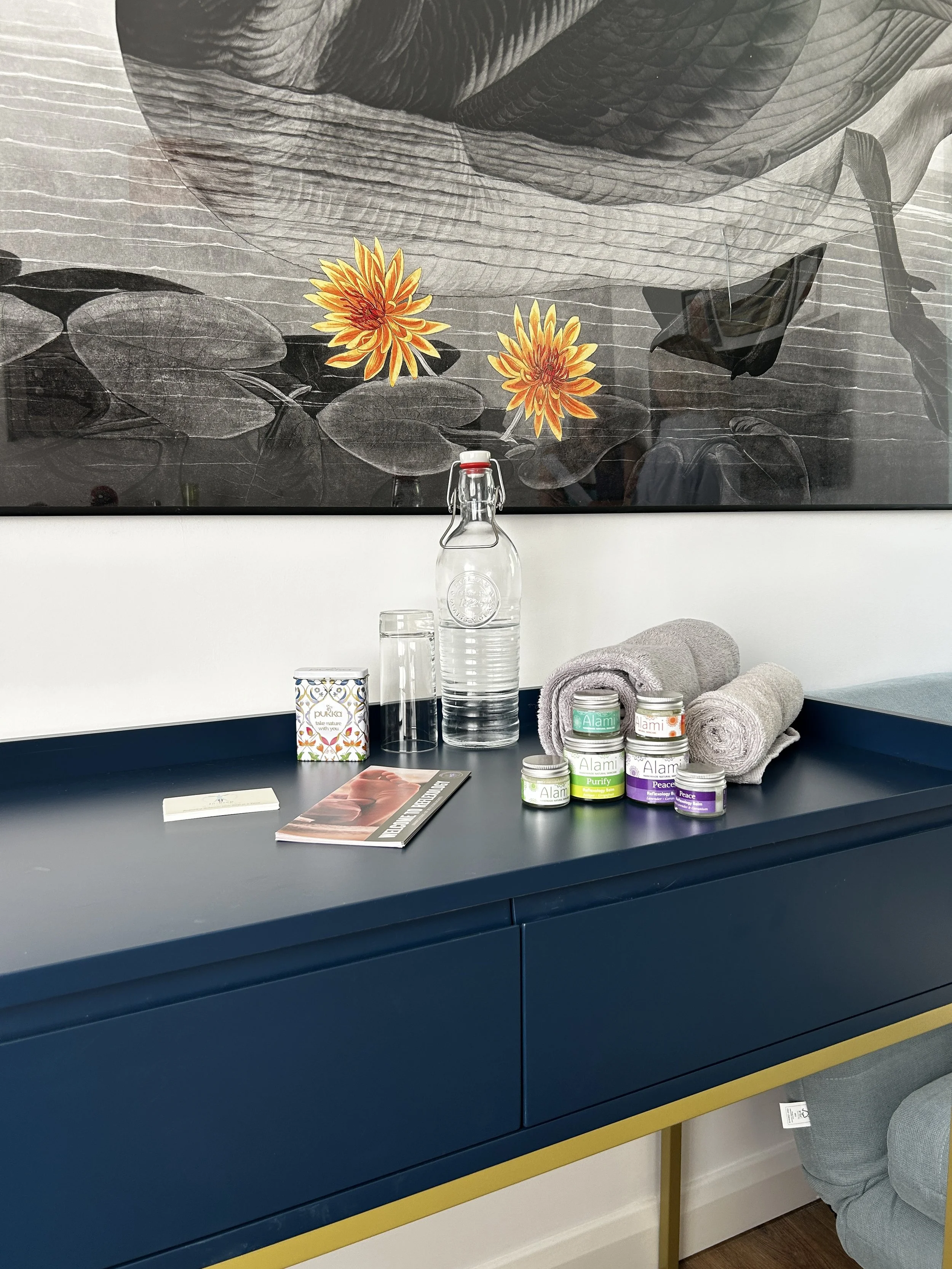 A blue and yellow sideboard with towels, jars of cream, a bottle of water, a glass, a box of tea, and a magazine on top. Behind, a large picture of a water lily with two orange and yellow flowers is on the wall.