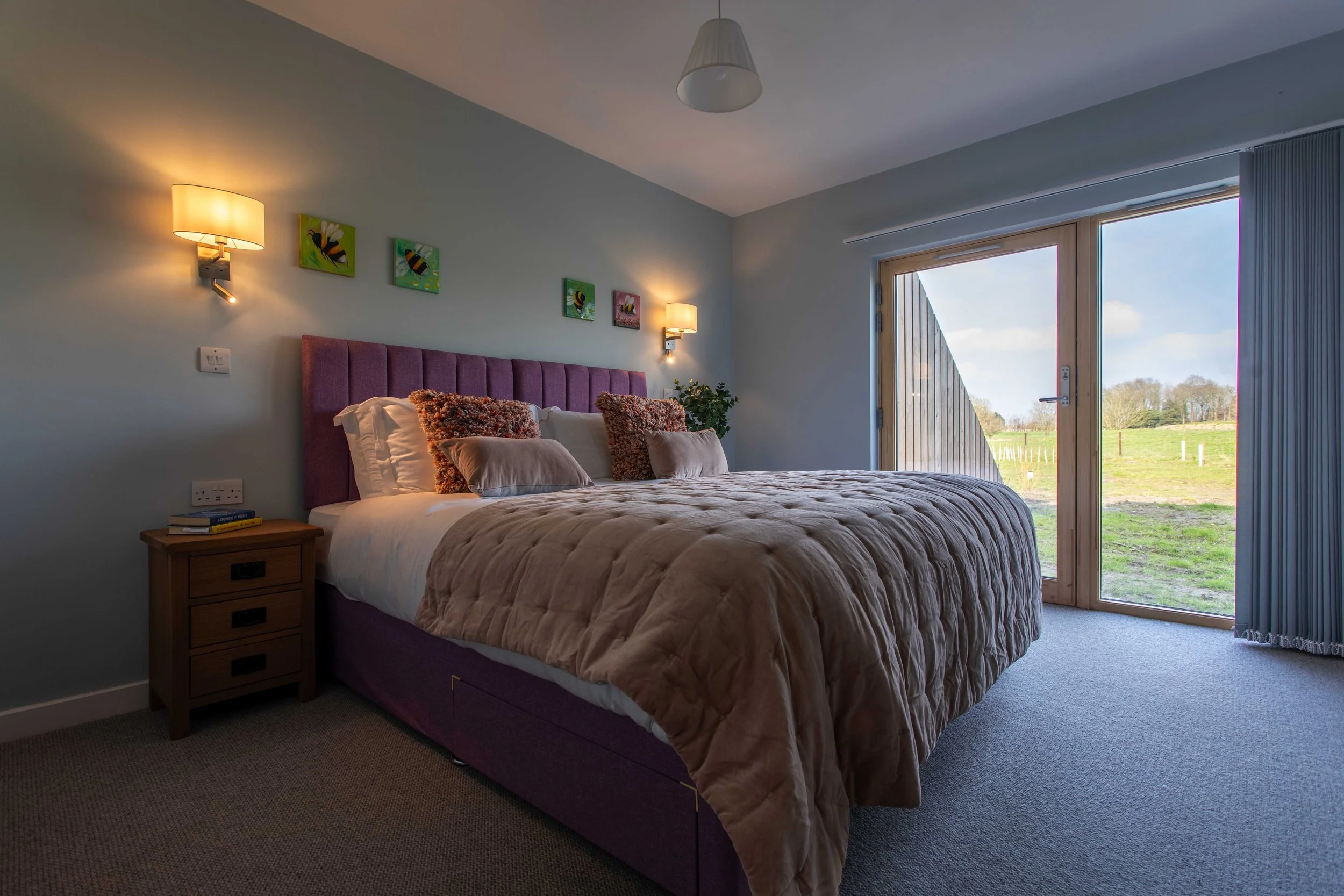 A bedroom with a purple upholstered bed, beige bedding, and multiple pillows including brown, beige, and white ones. There are two wall-mounted lamps and three small colorful paintings of bees above the bed. A wooden bedside table with books is on the left. Large glass door with blue curtains leads outside to a green field.