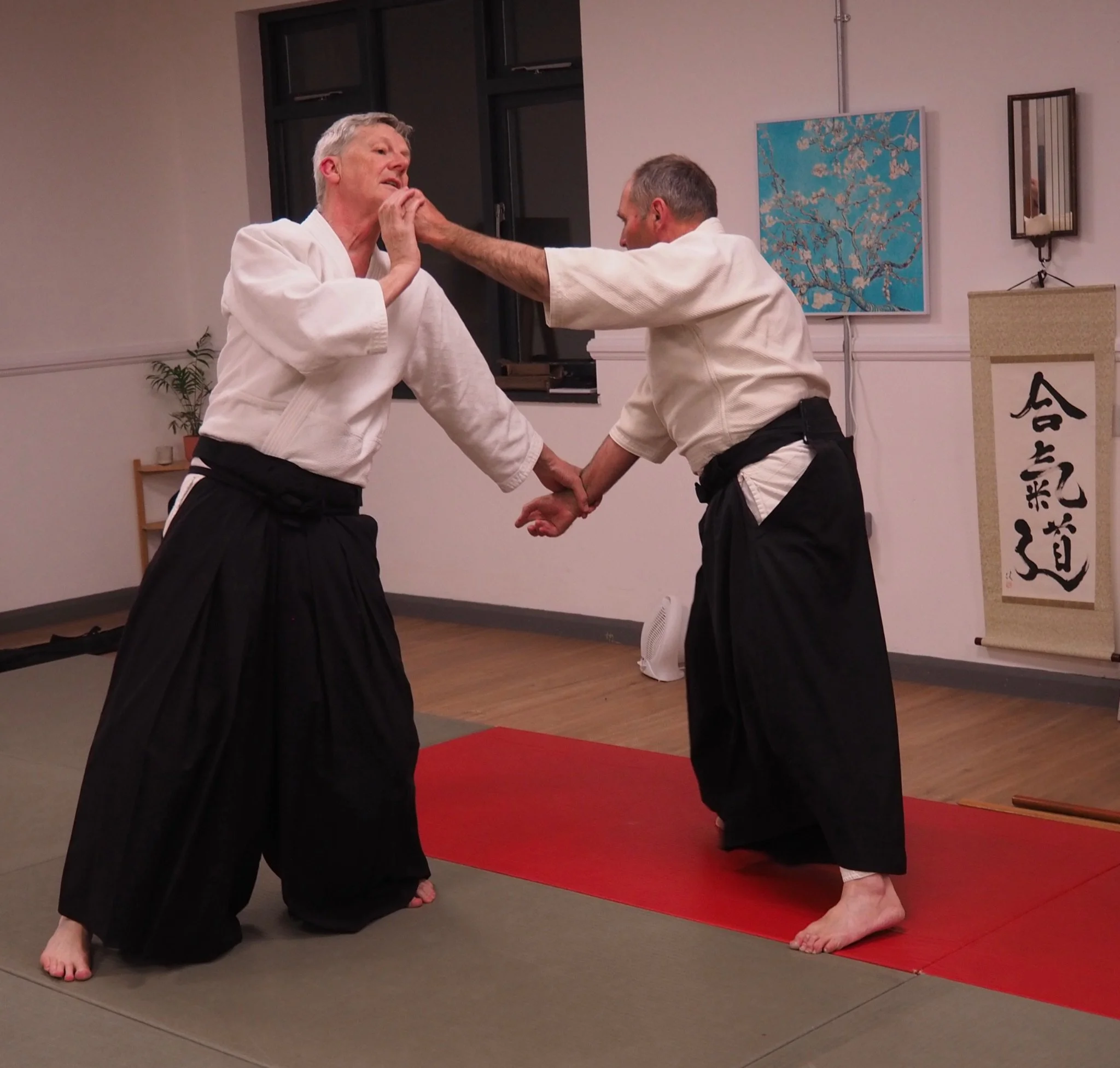 Two men practicing martial arts on a red mat indoors. Both wear white uniforms and black belts. One man is holding the other's face, and they are gripping each other's wrists. A painting of cherry blossoms and some decorative items are on the wall behind them.