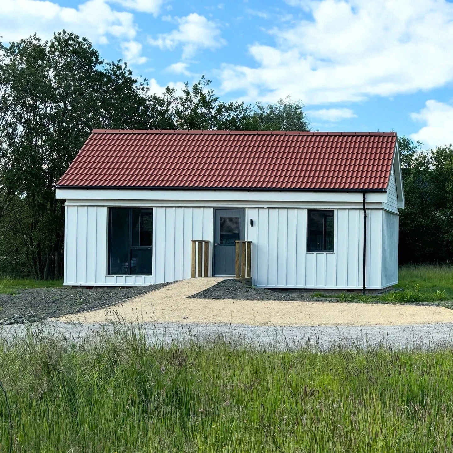 Small white house with black trim, red tiled roof, and a gravel driveway, surrounded by trees under a partly cloudy sky.