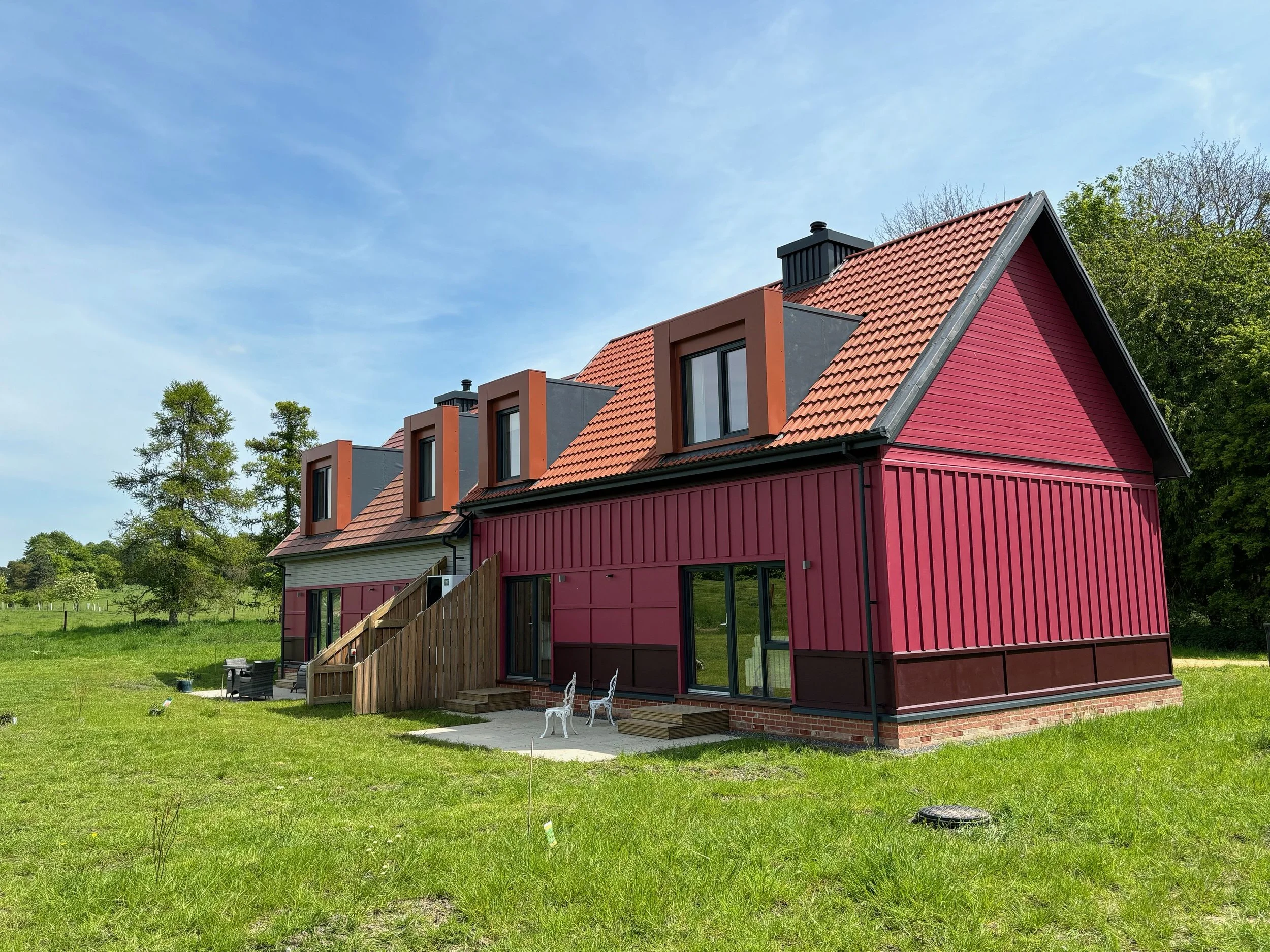 Modern two-story house with a red and gray exterior, multiple dome-shaped windows, set on a green grassy field beneath a blue sky with some wispy clouds.