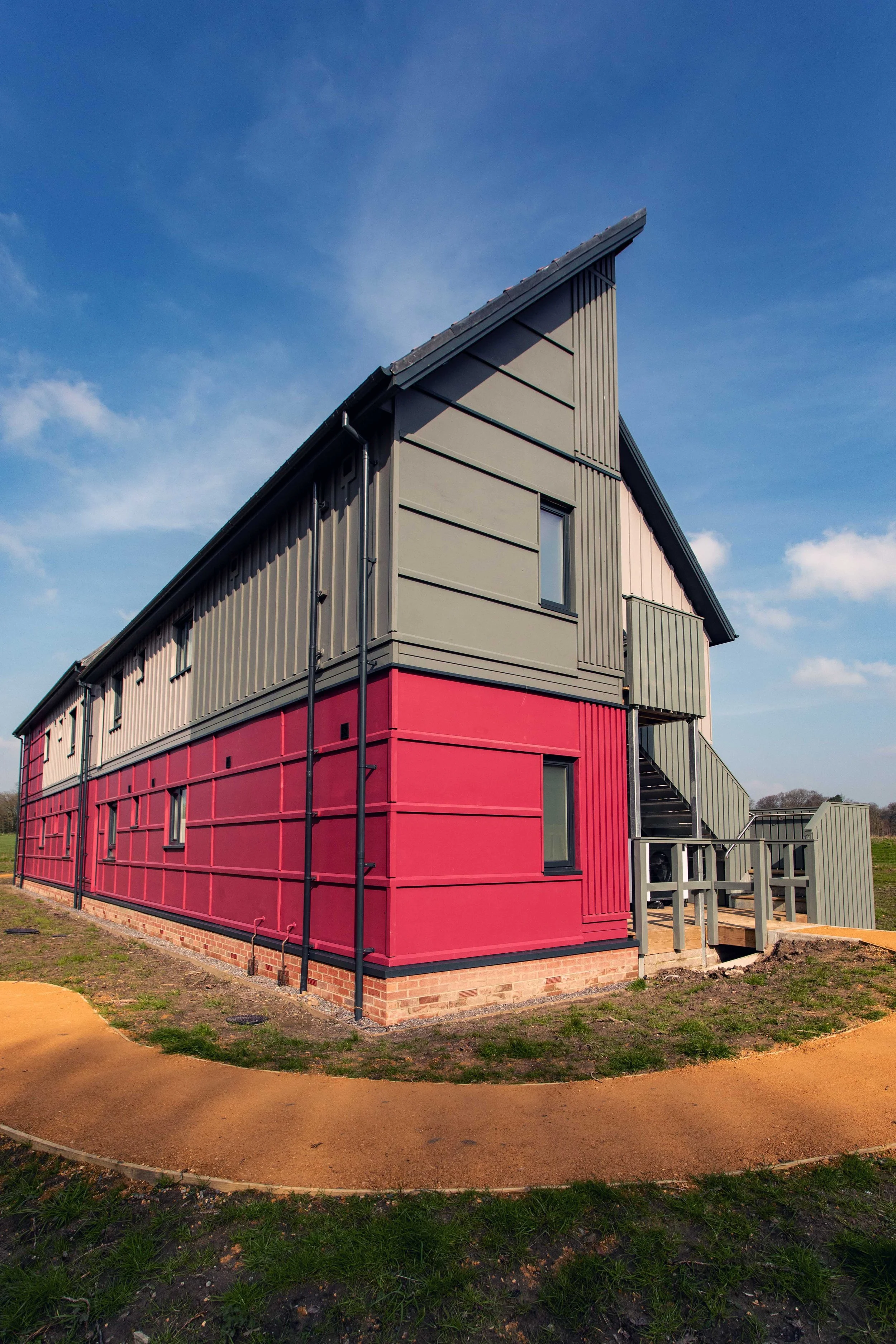 Modern building with red and gray exterior, multiple windows, and an external staircase, situated outdoors under a clear blue sky.