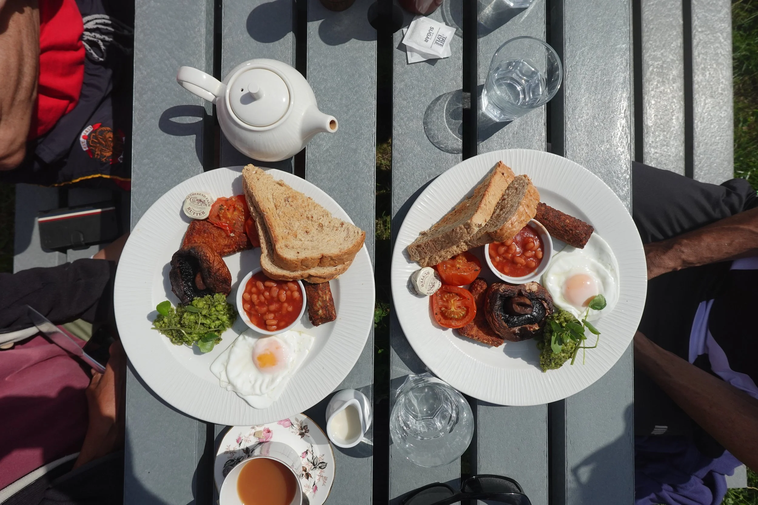 Two breakfast plates with fried eggs, baked beans, grilled tomatoes, mushrooms, toast, and grilled meat, set on a gray outdoor table with a glass of water, teapot, and condiments.