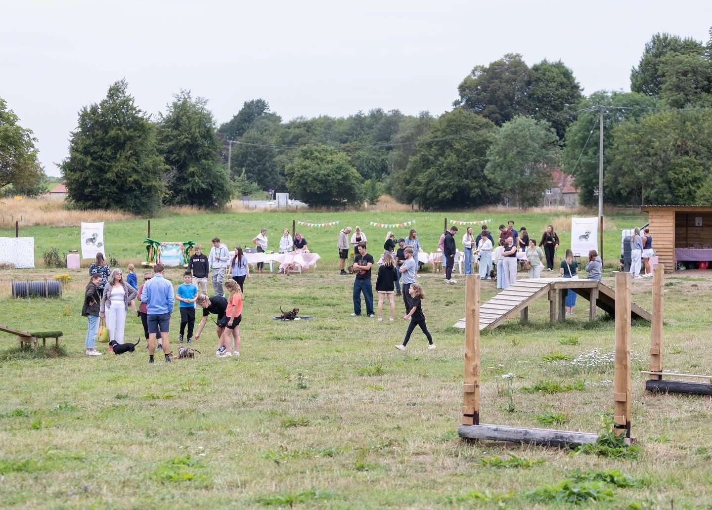 People gathered outdoors at a community event in a grassy field, with tables, banners, and a small wooden structure, surrounded by trees and open sky.