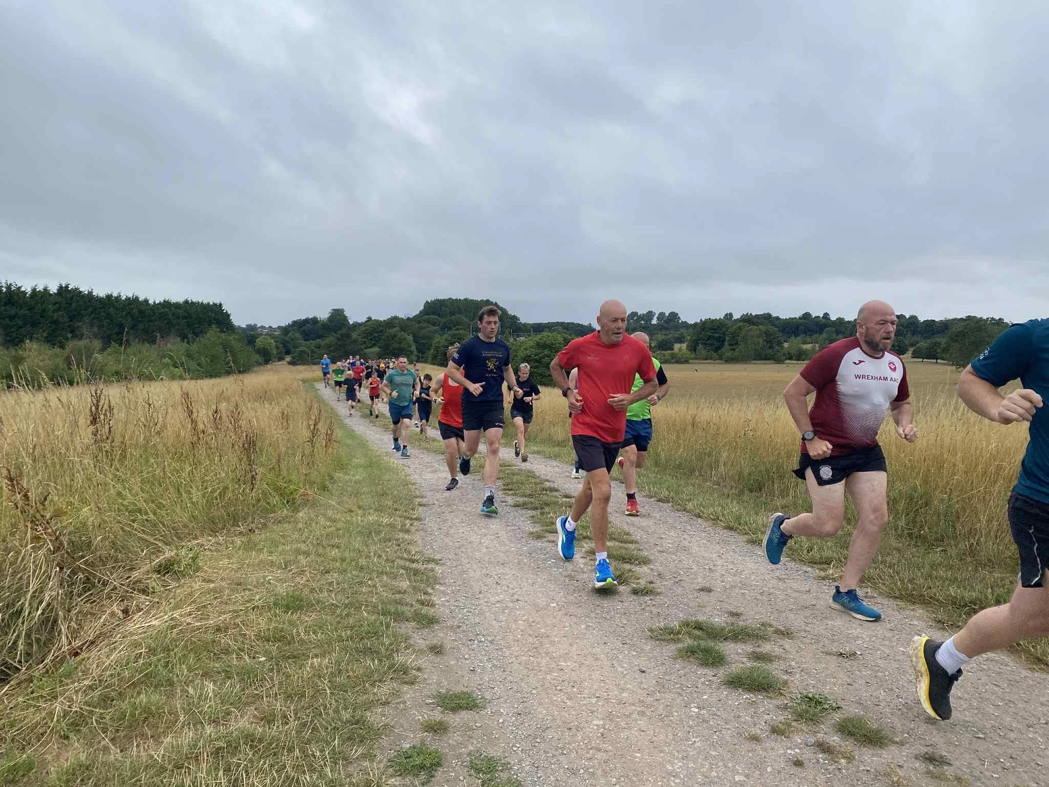 A group of people, including both men and women of various ages, participating in a running event along a rural dirt trail through a wheat field with trees and overcast sky in the background.