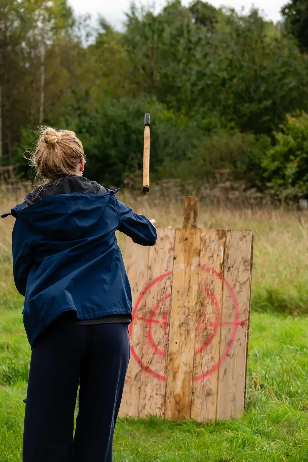 A woman throws an axe at a wooden target with a red target circle painted on it, outdoors on a grassy field with trees in the background.